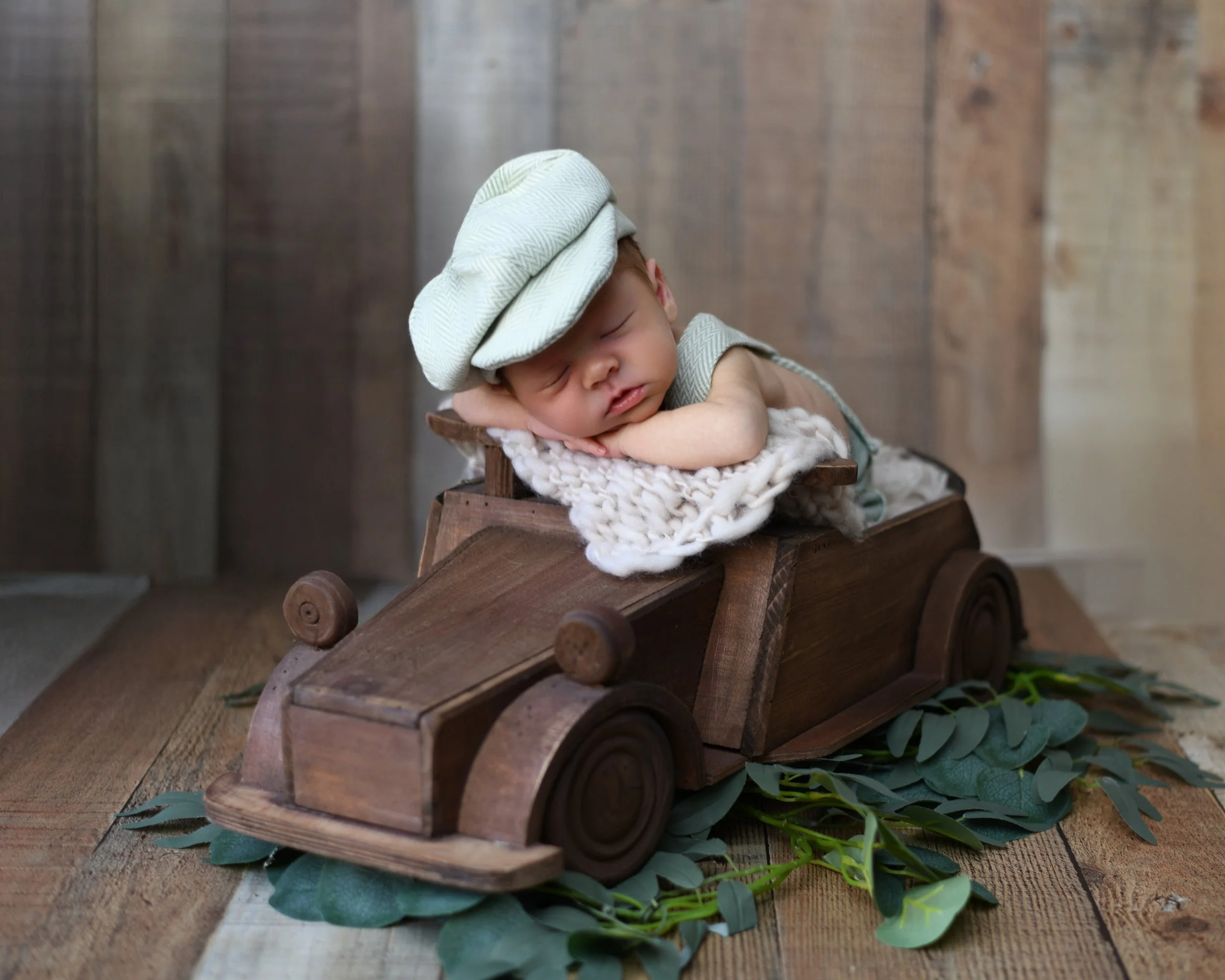 A sleeping baby in vintage clothing lies on a blanket inside a wooden toy car, wearing a light green cap, placed on a wooden floor with greenery underneath against a wooden wall background.