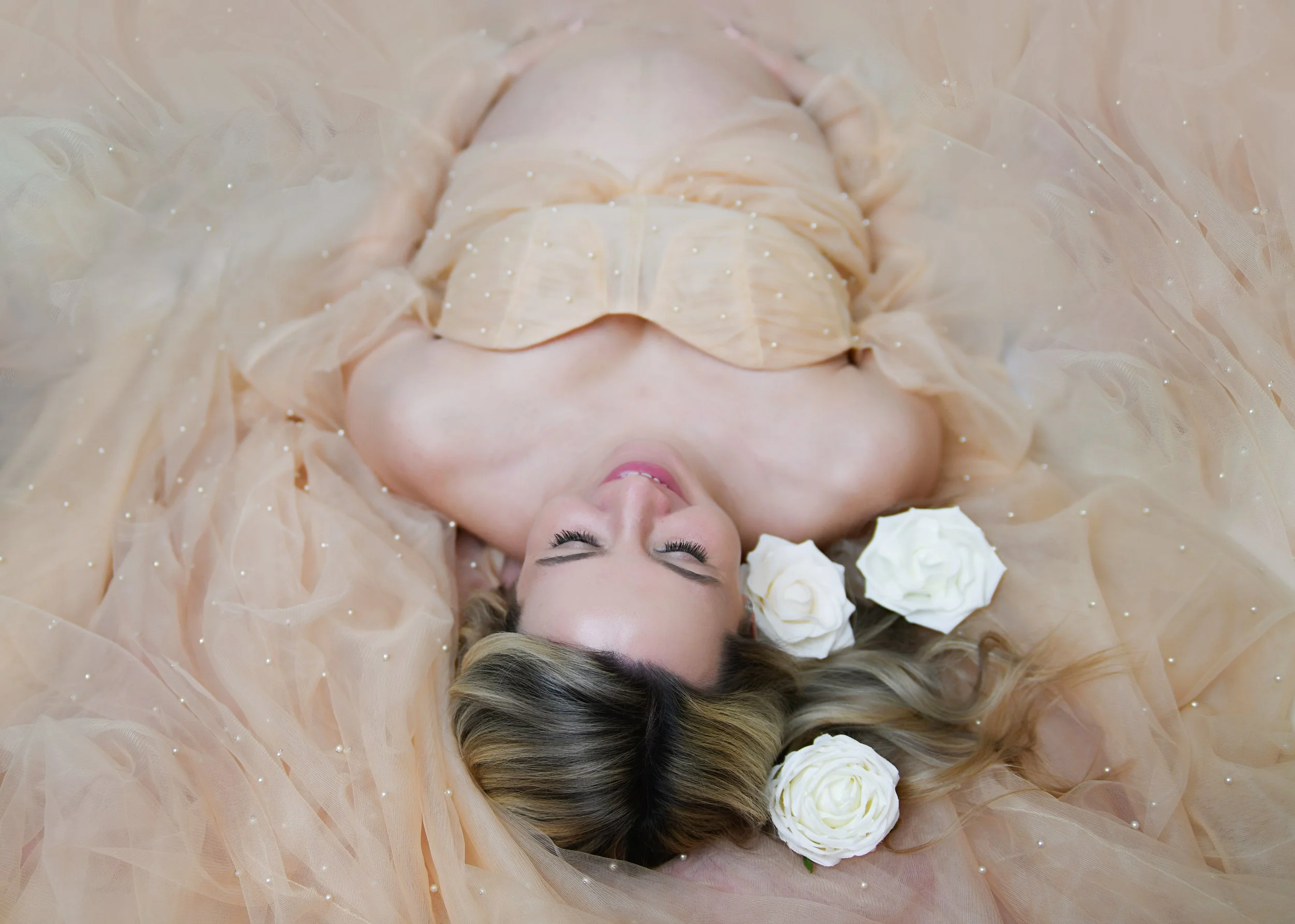 A woman with long blonde hair lays on a peach-colored tulle fabric, surrounded by white roses, with her eyes closed and a peaceful expression.