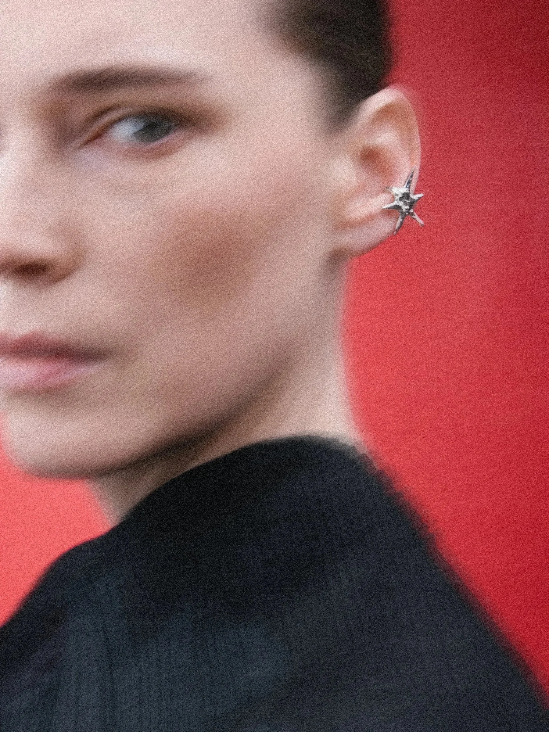 Close-up of a young woman wearing a star-shaped silver earring, and a black top, against a red background.