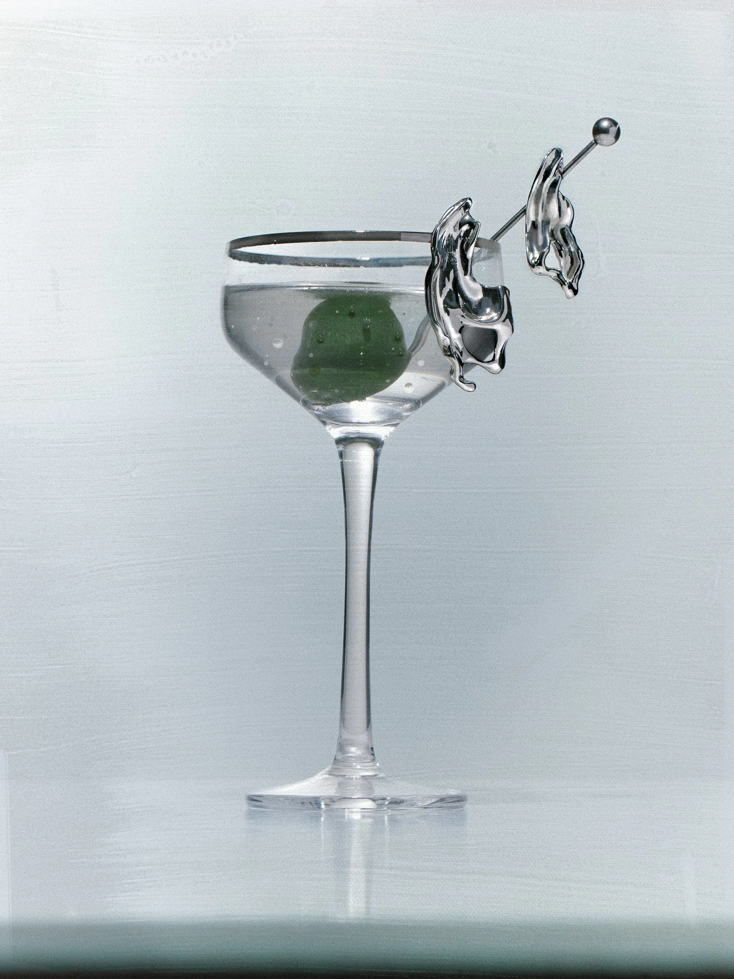 A martini glass filled with a clear liquid, garnished with a green olive and a metallic cocktail pick with sterling silver wavy mismatched earrings, against a plain light background.