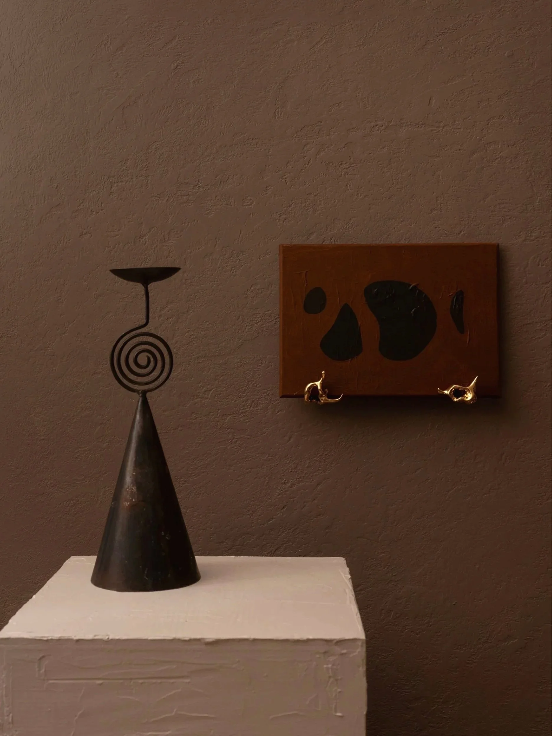Decorative sculpture placed on a white pedestal, with a dark abstract painting with gold sculptural organic shaped art ledge on a brown wall in the background.