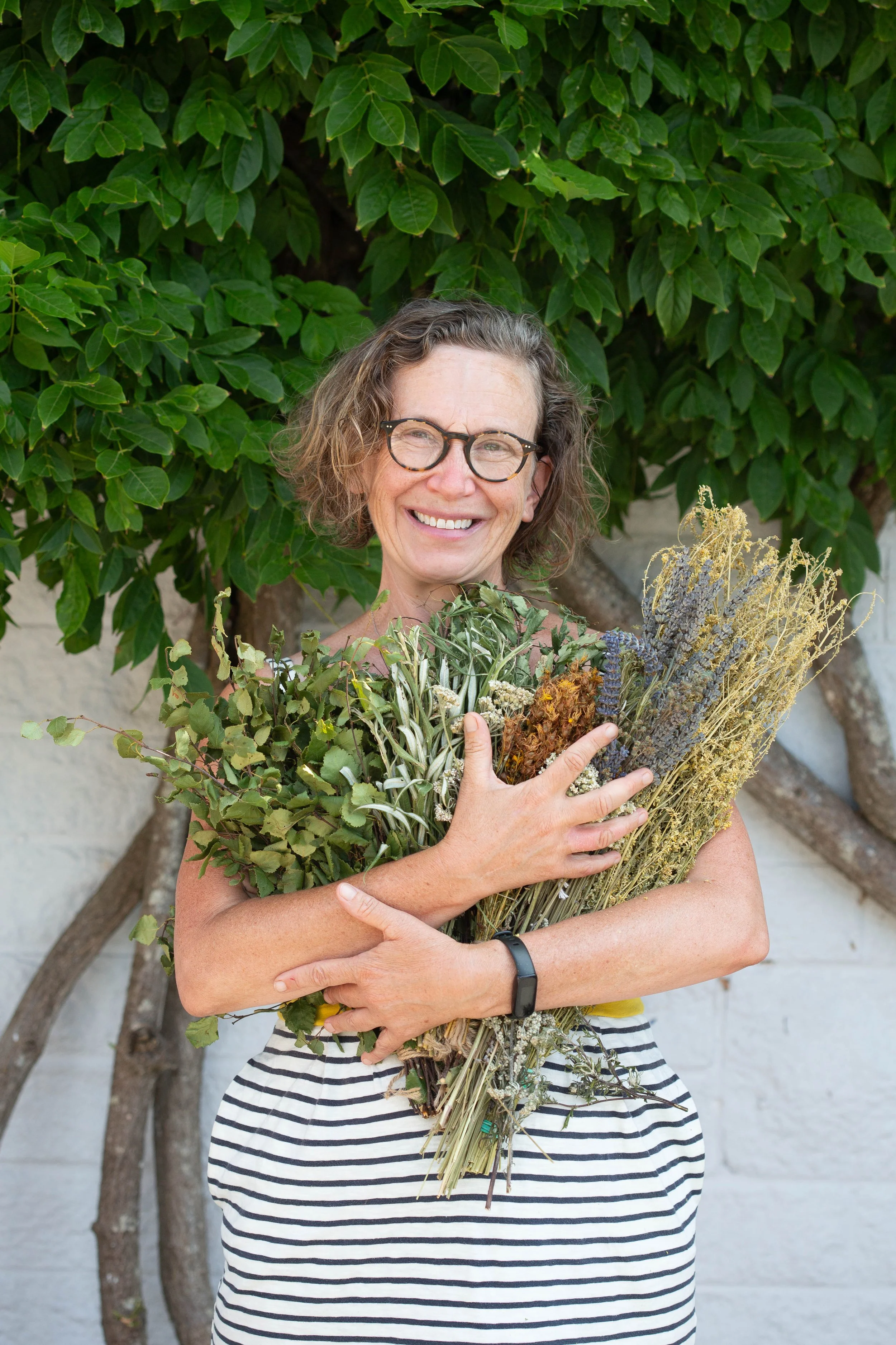 A woman with curly hair, glasses, and a striped dress holding a large bouquet of dried and fresh herbs and plants, standing outdoors in front of a leafy green bush.