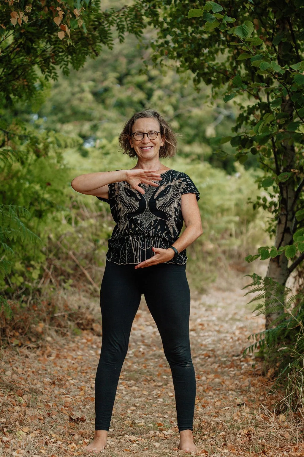 A woman practicing yoga outdoors on a wooded trail, standing with one hand above her head and the other resting on her abdomen, smiling at the camera.
