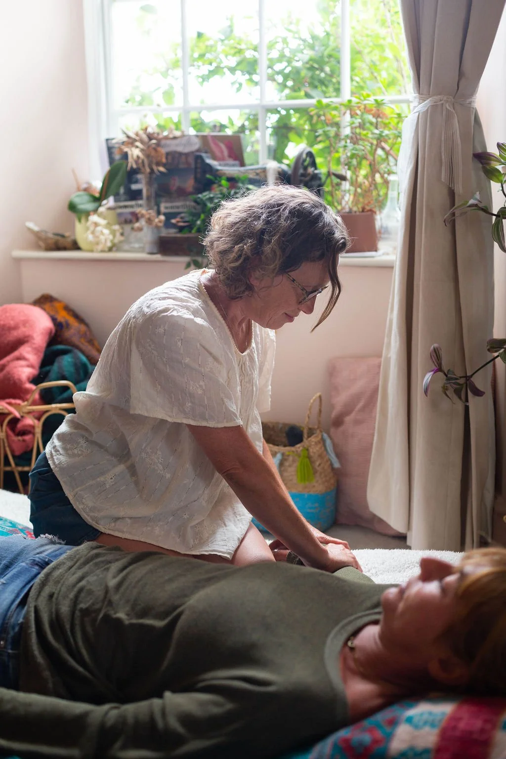 A woman providing CPR to a person lying on the floor inside a well-lit room with large window and plants.