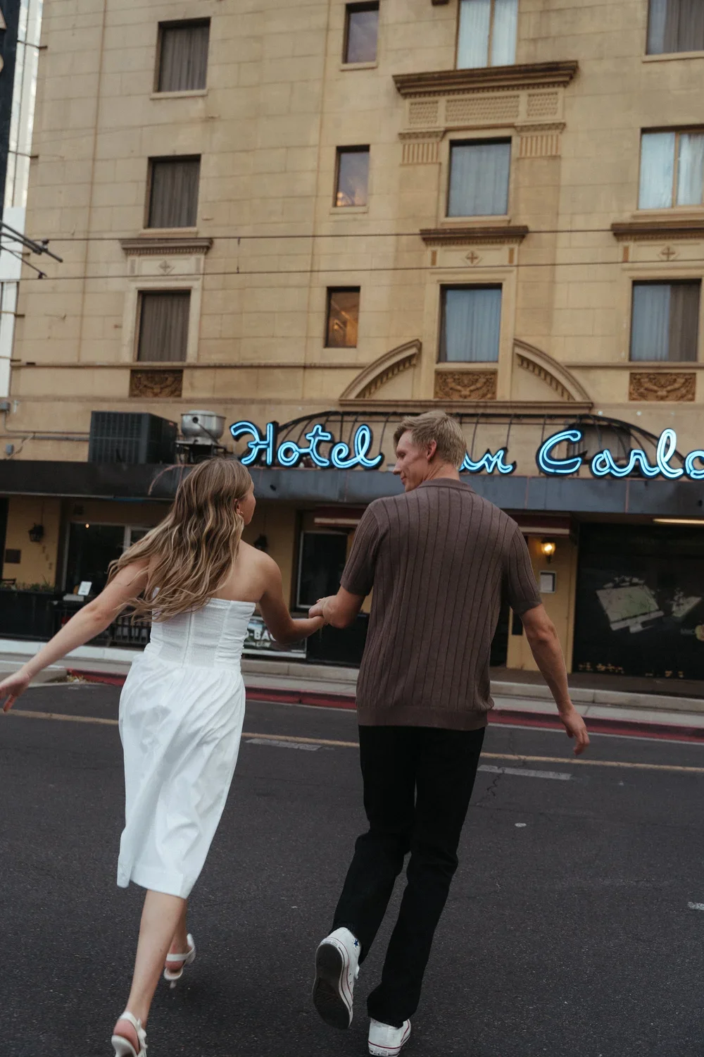 A couple holding hands and running on the street in front of a hotel with a neon sign that reads 'Hotel um Carlso' in an urban area.