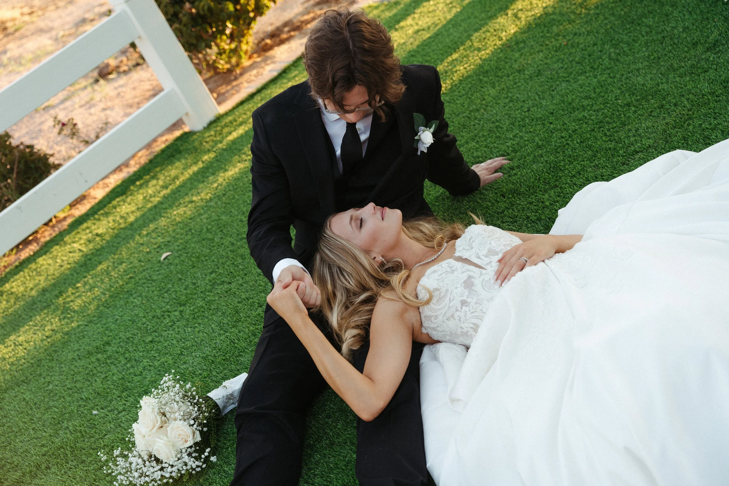 A groom and bride lying on the grass during their wedding, holding hands, with flowers in the background.