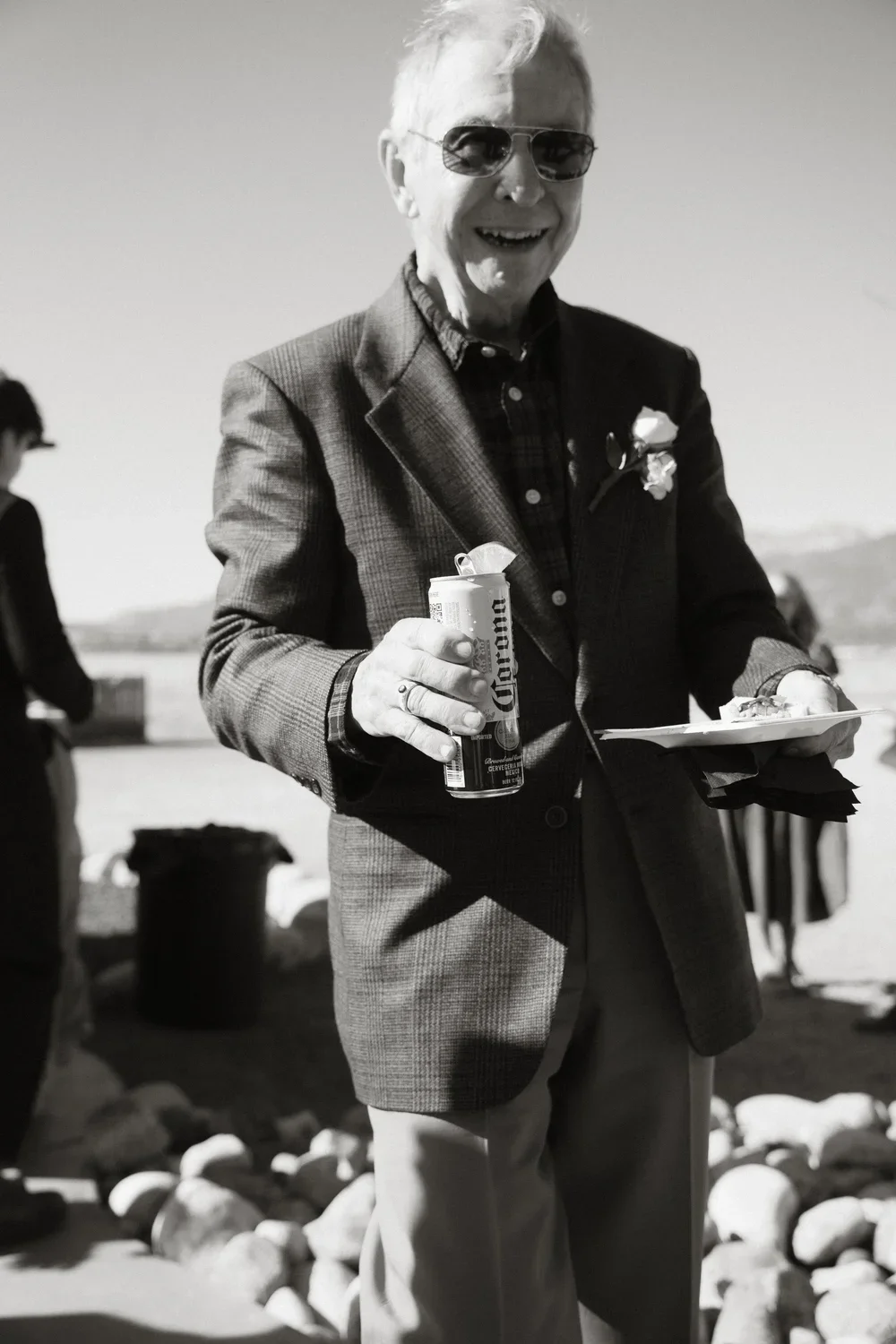 An elderly man smiling, wearing sunglasses and a checked blazer, holding a can of Corona beer in one hand and a plate of food in the other, outdoors with people and landscape in the background.