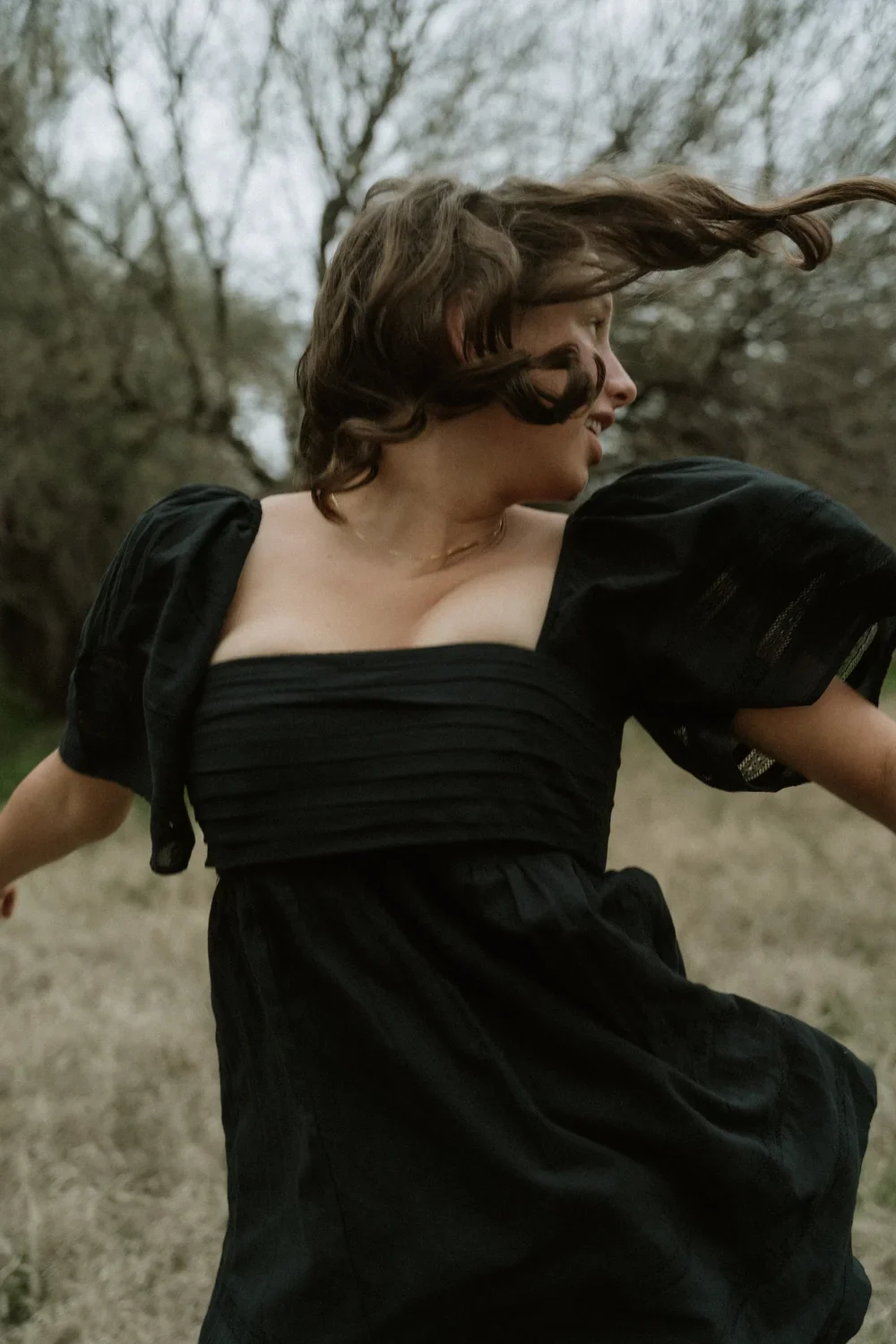 A woman in a black dress is outdoors, with wind blowing her hair, turning her face to the side and smiling.