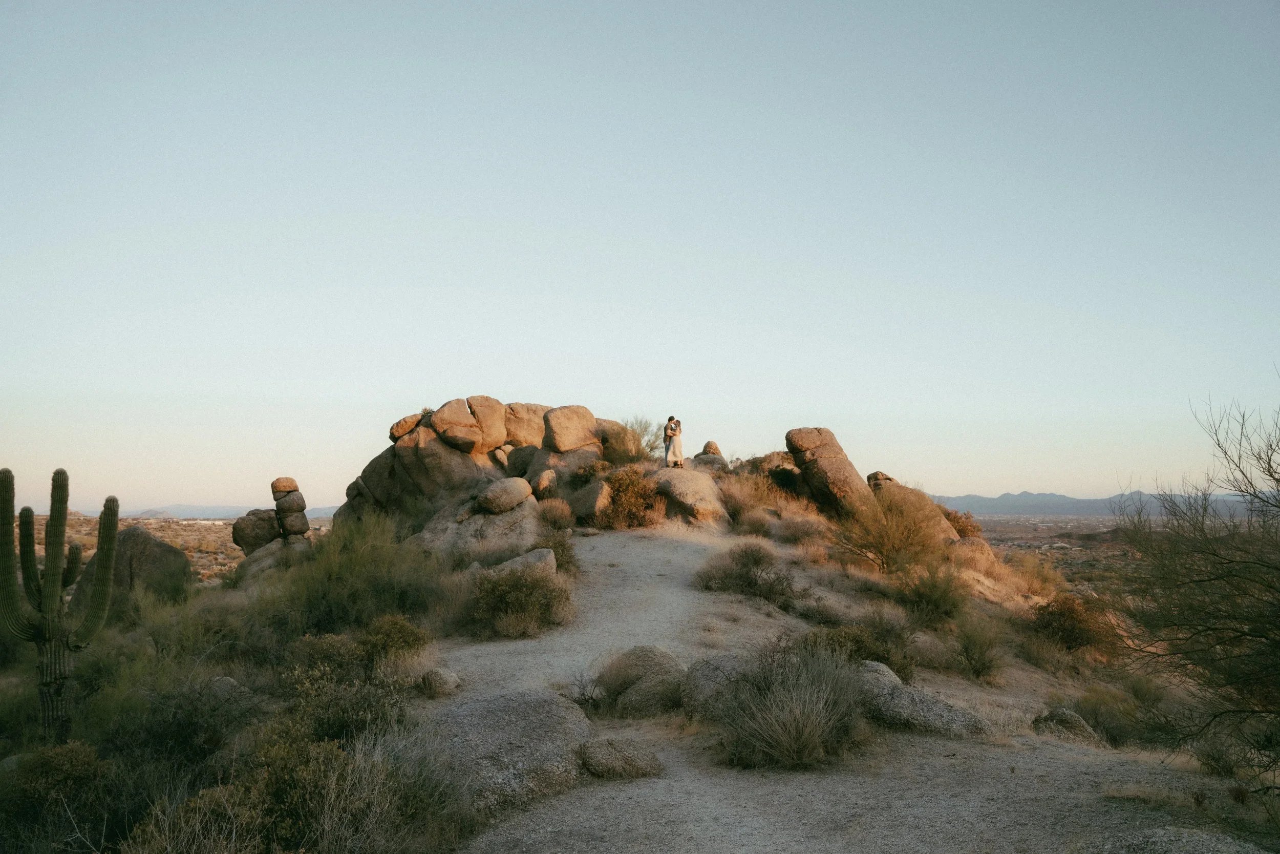 A desert landscape with large rock formations, cacti, shrubs, and a dirt path. Two people are standing on a rocky hilltop, taking a photo together. The background features distant mountains and a clear sky.