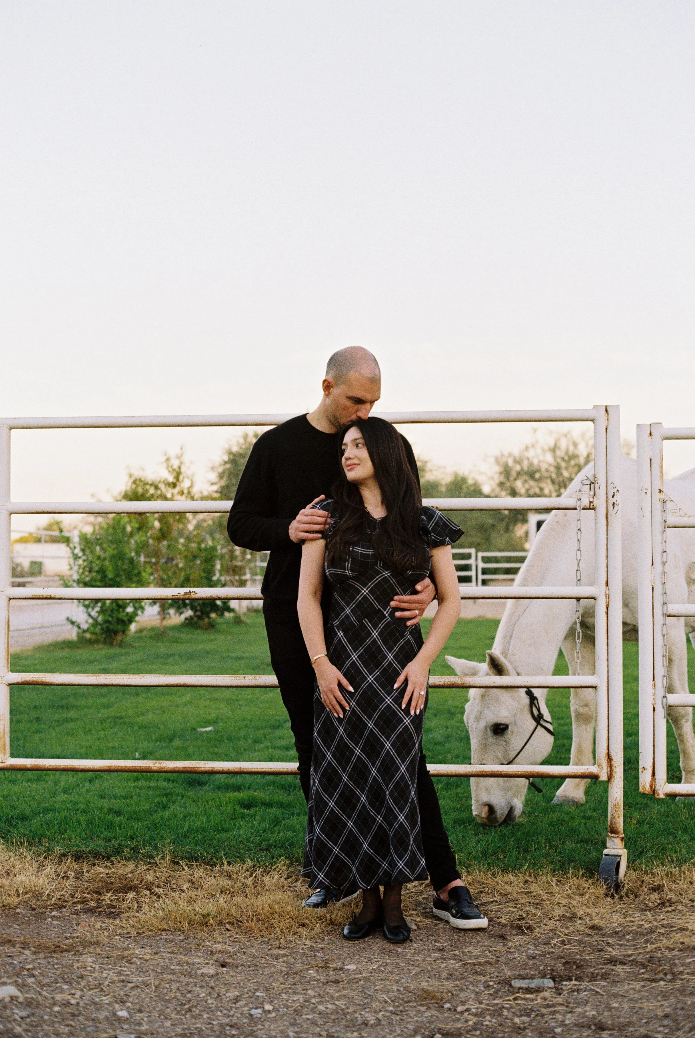 A man and a woman standing together in front of a white horse at a farm or ranch, with green grass, trees, and a fence in the background during sunset.