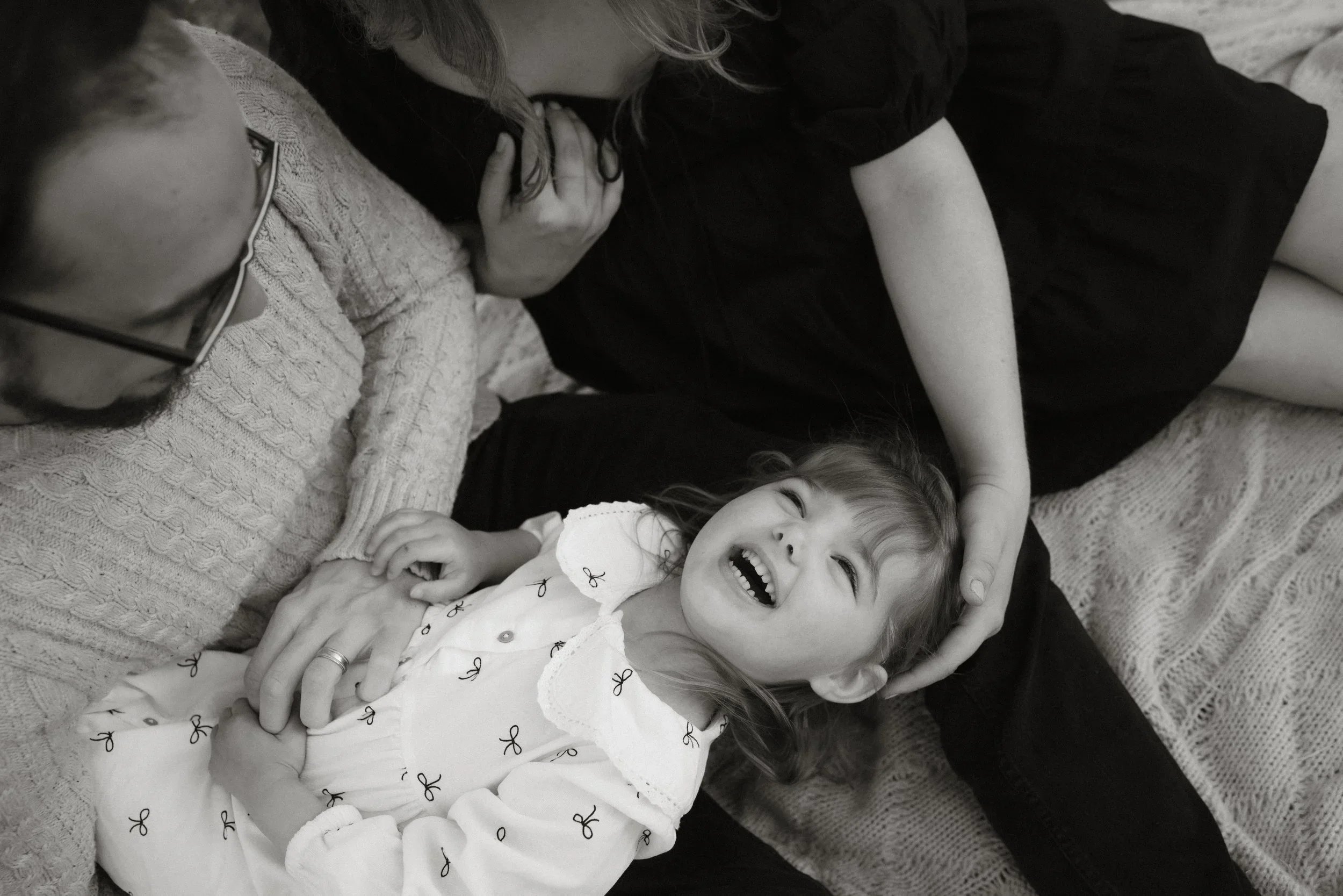 A black and white photo of two women and a young girl lying on a blanket. The women are cuddling with the girl, who is smiling and laughing with her eyes closed. One woman wears glasses and a cozy sweater, while the other wears a dark top. The girl is wearing a light-colored shirt with a bow pattern and a collar.