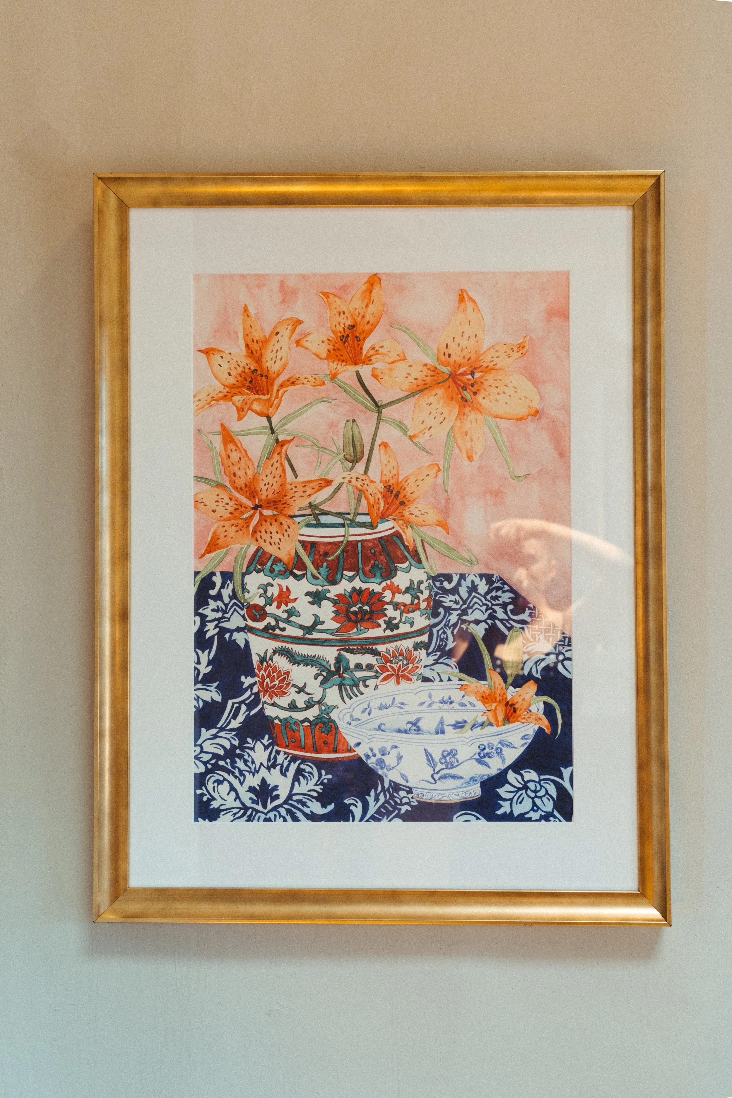 Framed artwork of orange lilies in a decorative vase, with a bowl of matching flowers, on a patterned blue tablecloth.