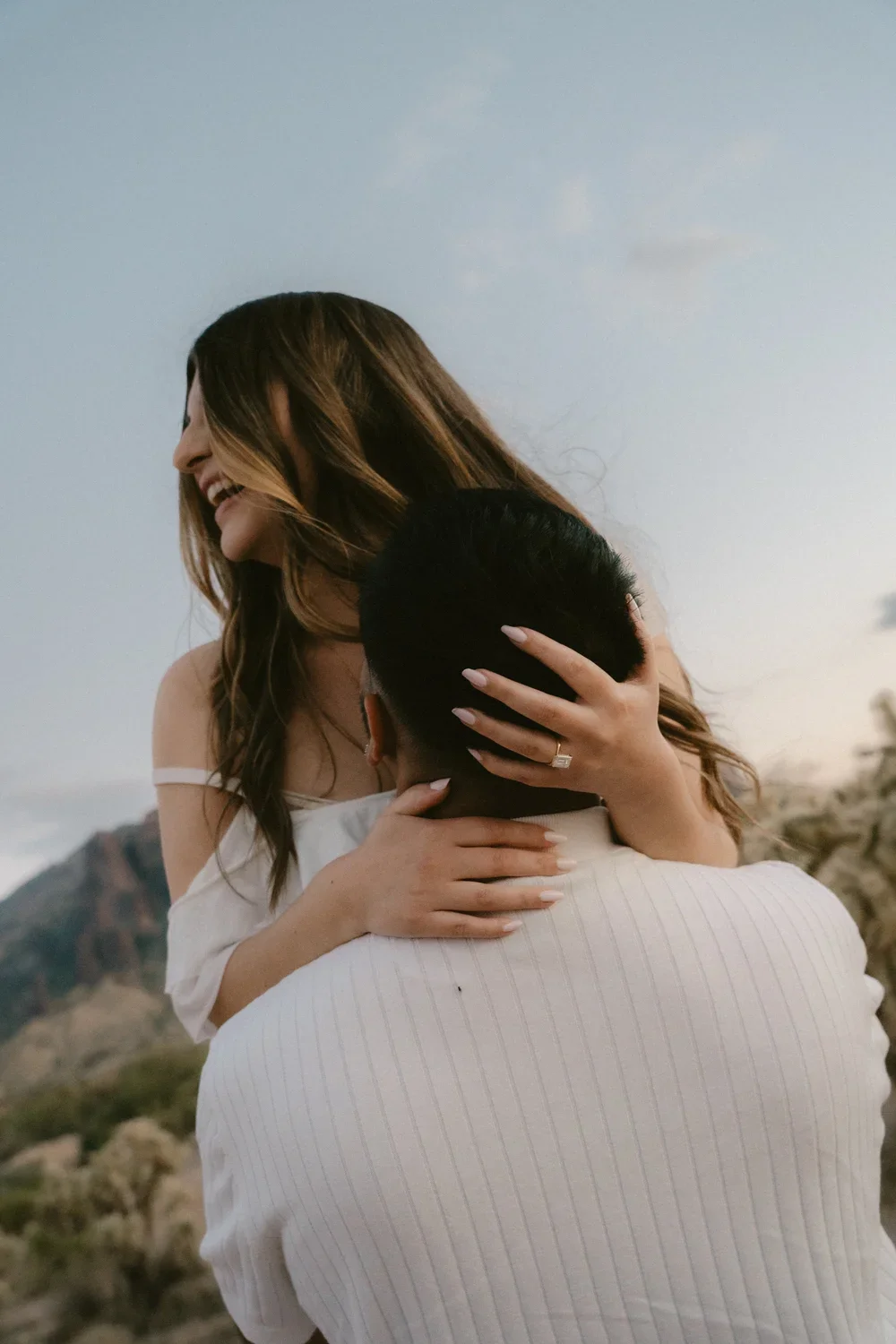 A woman hugging a man outdoors, smiling with her eyes closed, mountain and sky in the background.
