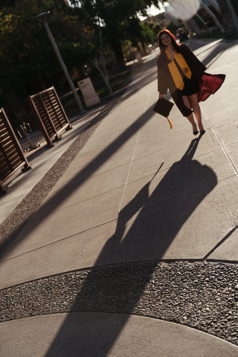 A woman in graduation attire walking on a sidewalk, holding a diploma and a graduation cap, with the sun casting a long shadow behind her.