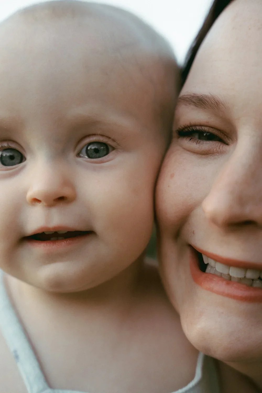 Close-up of a smiling woman with a partly closed eye next to a curious baby with blue eyes, both showing their faces close together.