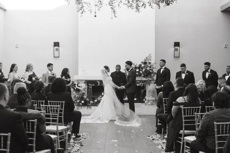 A wedding ceremony indoors with a bride and groom holding hands, surrounded by their wedding party and seated guests.