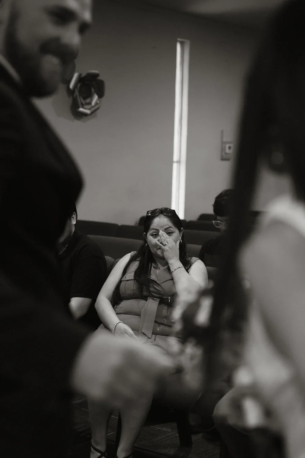 A woman sitting in a waiting room, covering her nose or mouth with her hand, surrounded by people and framed by blurred figures in the foreground.