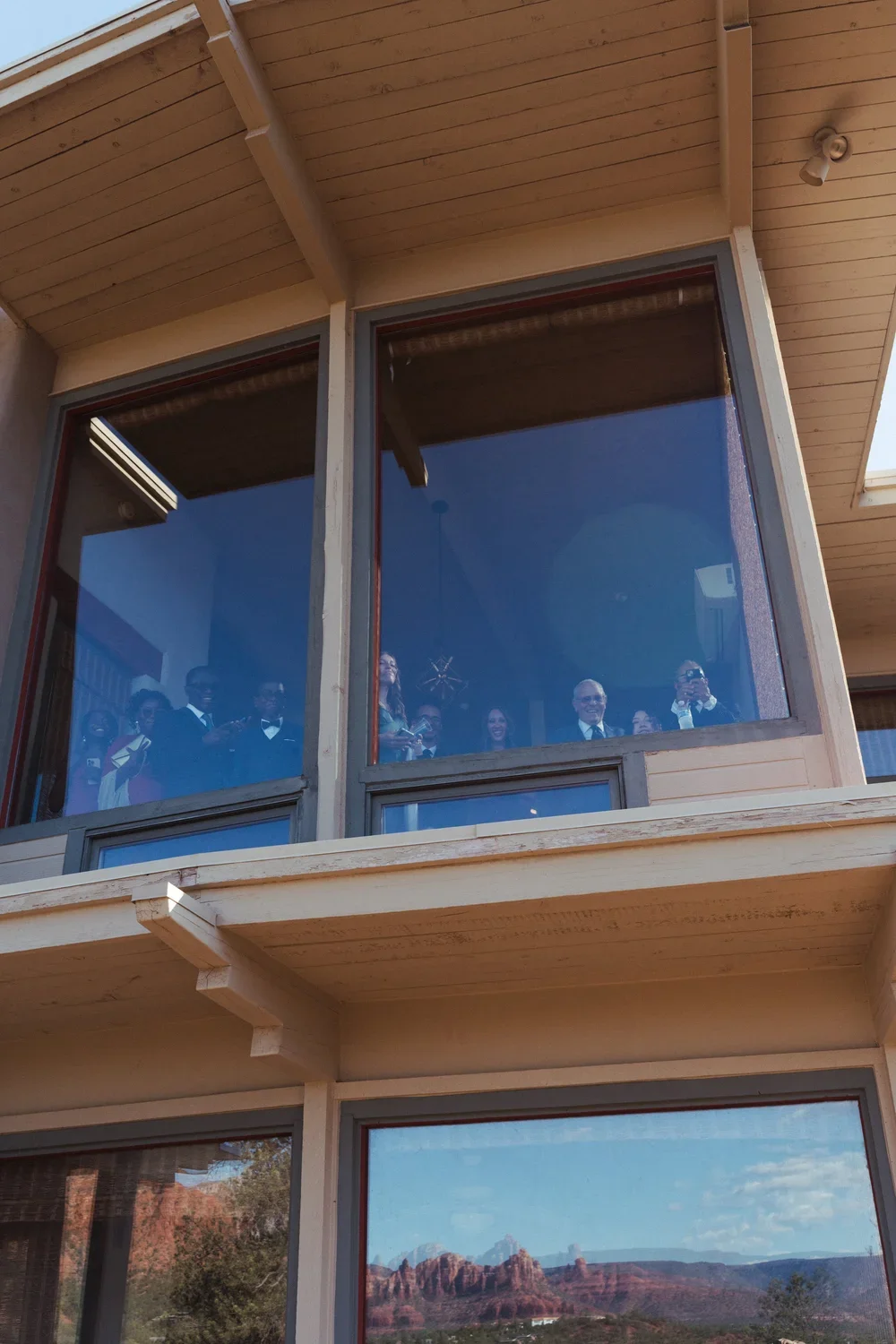 People gathered inside a building, viewed through large glass windows, with a scenic desert landscape with red rock formations visible outside.