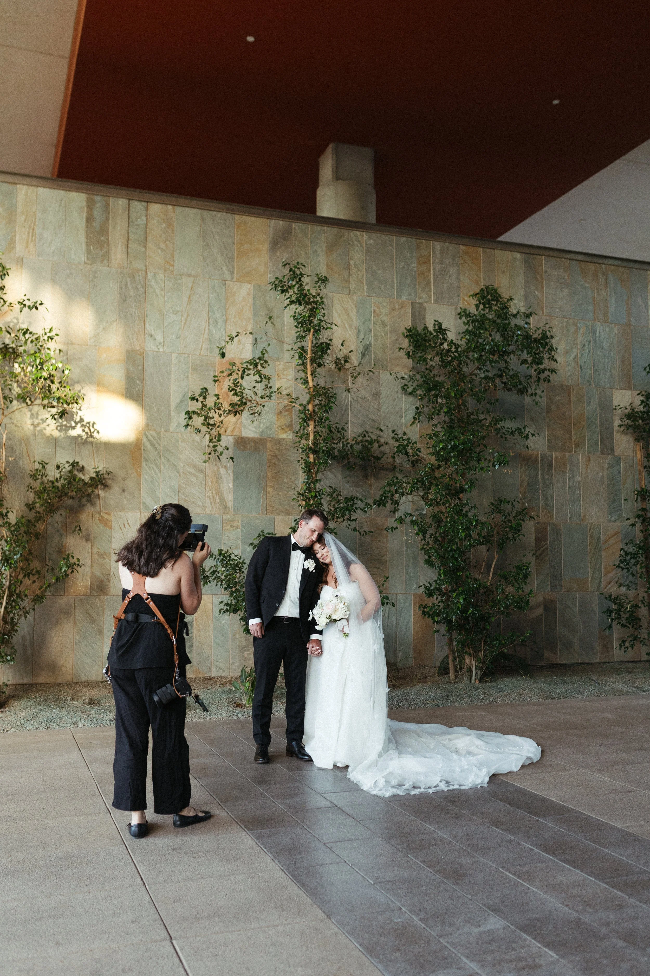 A bride and groom hold hands and pose for a wedding photo while a photographer takes their picture against a stone wall and plants indoors.