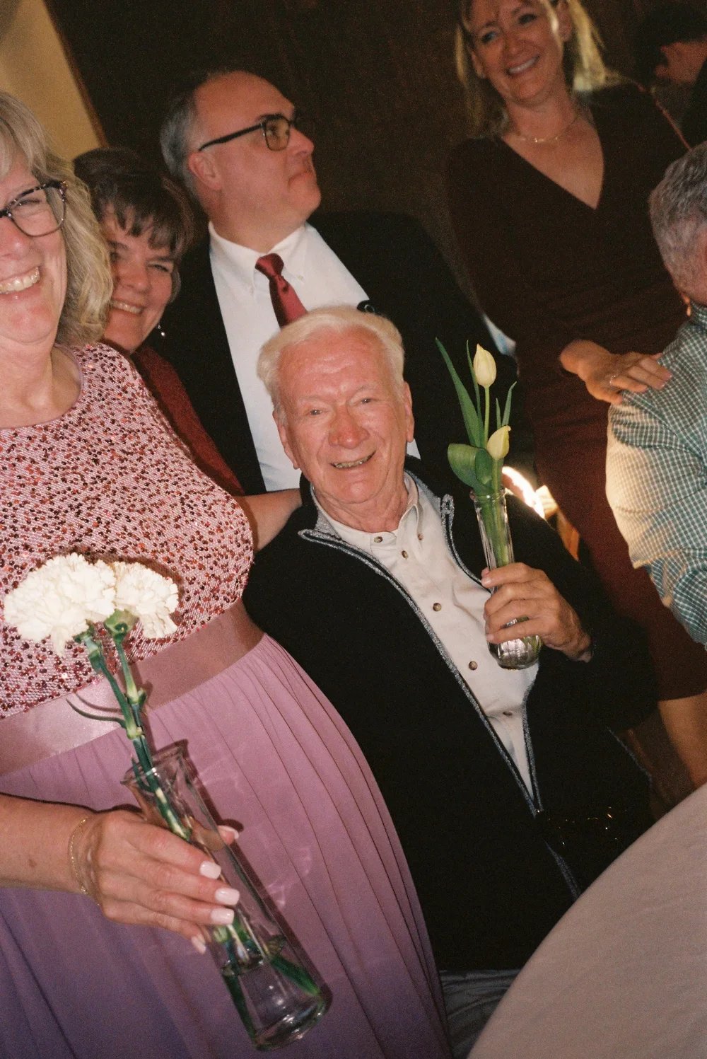 Group of smiling elderly and middle-aged people at a celebration, holding small vases with white flowers, gathered around a table.