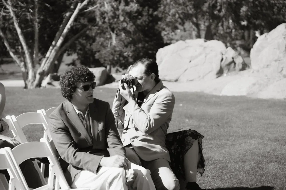 Two women sitting on white chairs outdoors. One woman with curly hair and sunglasses. The other woman is taking a photo with a camera, wearing a suit and skirt. Large rocks and trees in the background.