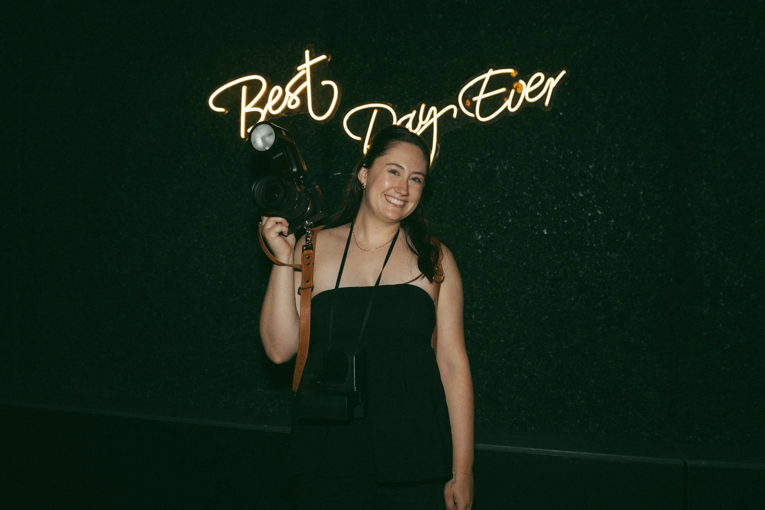 A woman smiling and holding a camera in her right hand, standing in front of a neon sign that reads "Best Day Ever" on a dark, speckled wall.