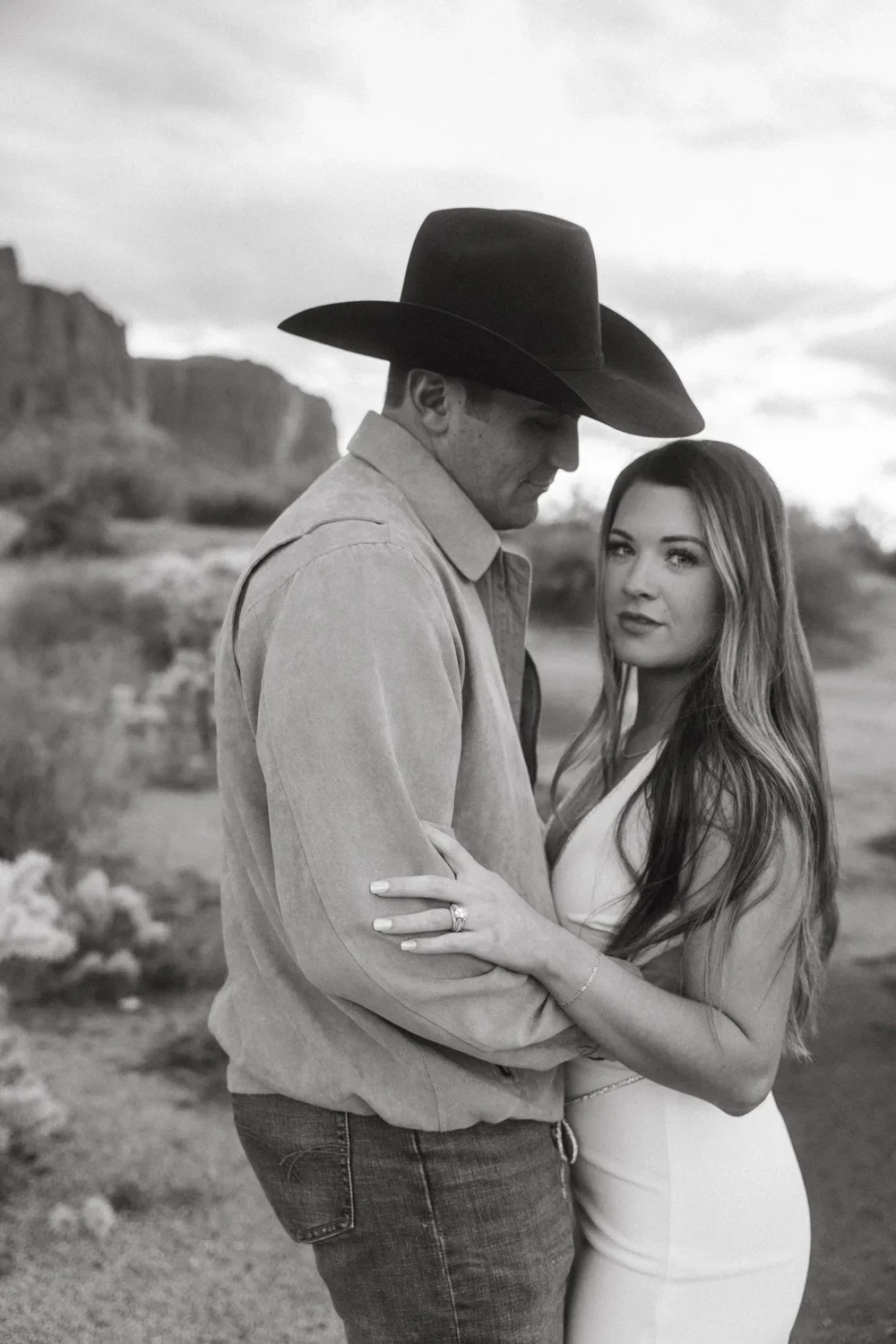 A black and white photo of a man and woman embracing outdoors in a desert landscape. The man is wearing a cowboy hat and a button-up shirt, while the woman has long hair and is dressed in a sleeveless dress. The woman is wearing an engagement ring, and they are looking at the camera.