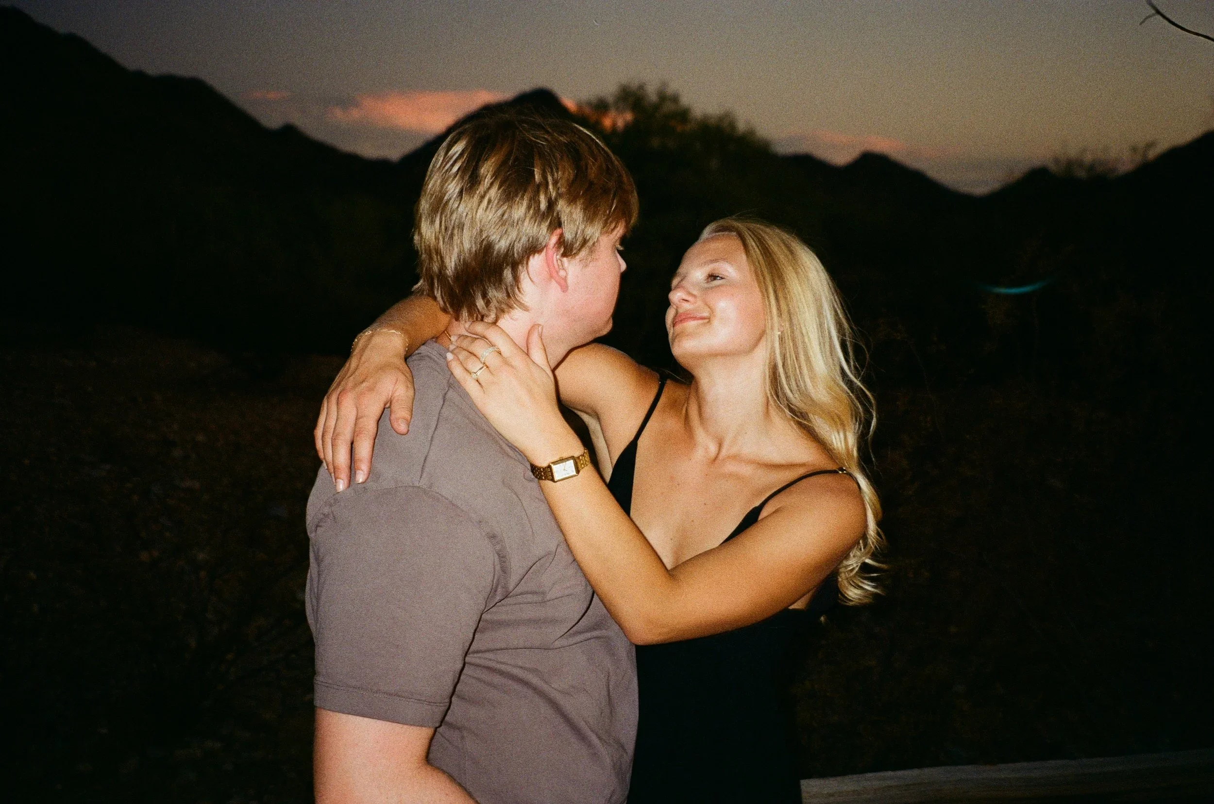 A couple embracing outdoors during sunset, with mountains and a partly cloudy sky in the background.