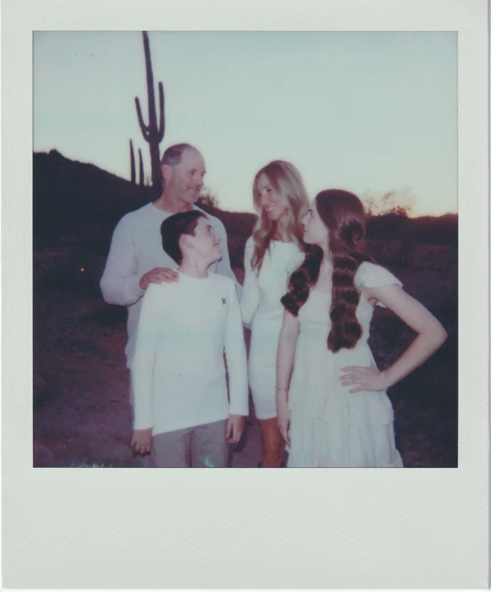 Four people, a man with gray hair, a woman with long blonde and a young girl with long brown hair, and a boy with short dark hair, standing in front of desert landscape with Saguaro cacti at sunset, smiling and engaging with each other.