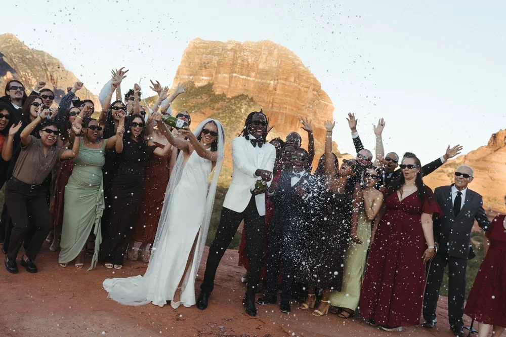 A wedding celebration with a bride and groom surrounded by friends and family outdoors near rock formations, with some people throwing champagne.