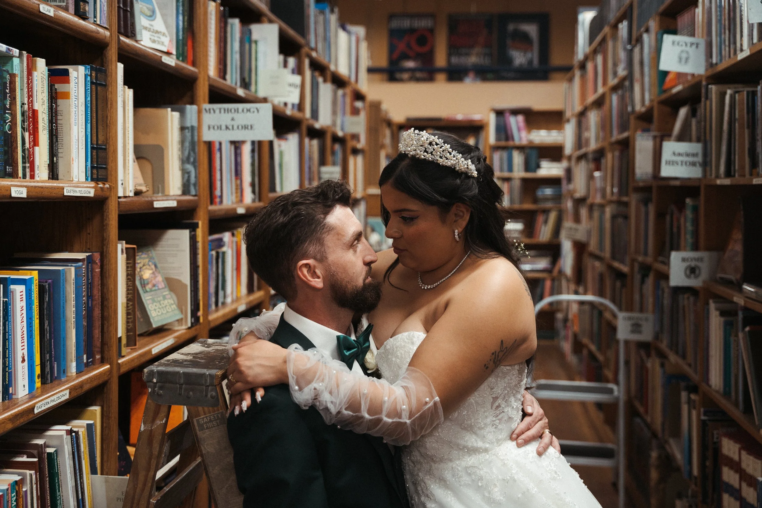 A bride and groom inside a library, the bride wearing a wedding dress with a pearl headband and jewelry, and the groom dressed in a tuxedo with a bow tie, embracing and looking at each other.