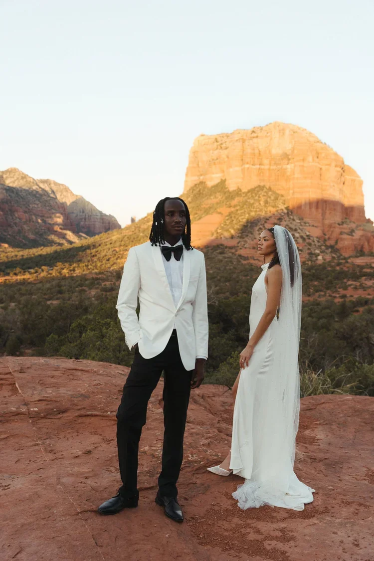 A wedding couple standing outdoors on a red rock terrain with a large canyon or mesa in the background during sunset, wearing formal wedding attire.