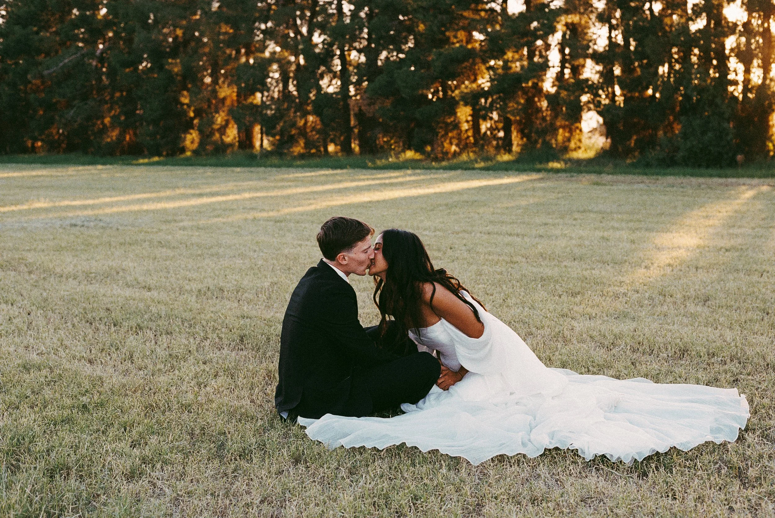 A bride and groom sitting on a grassy field kissing, with trees and sunset in the background.