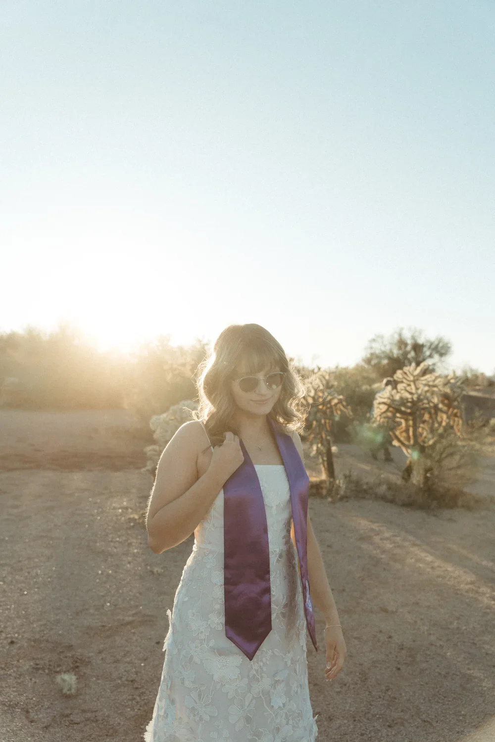 A young woman in a white floral dress with a purple graduation stole standing outdoors in a desert landscape at sunset, wearing sunglasses and carrying a small backpack.