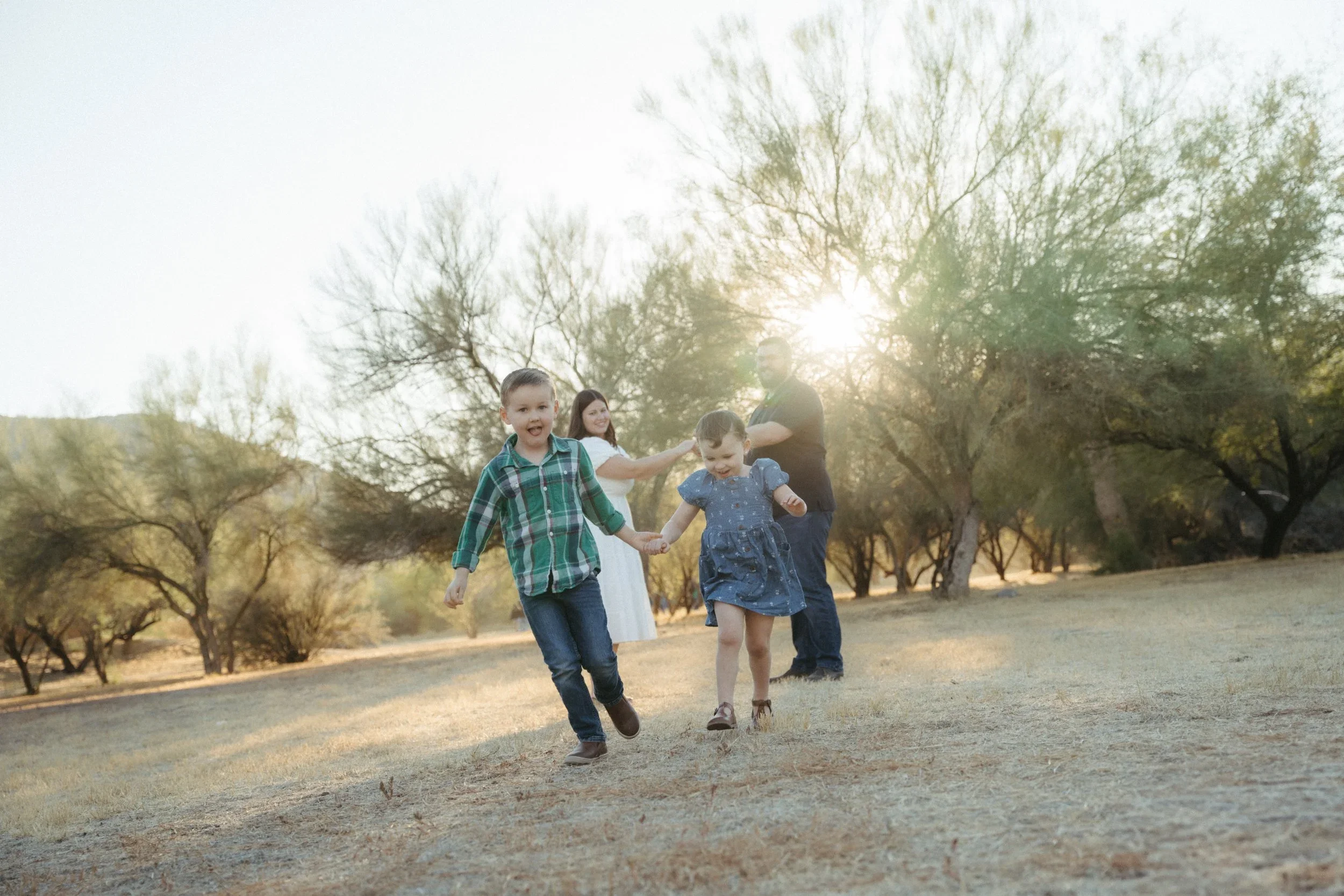 A family of five, including two young children and two adults, joyfully walk and play together outdoors during sunset in a park with trees.