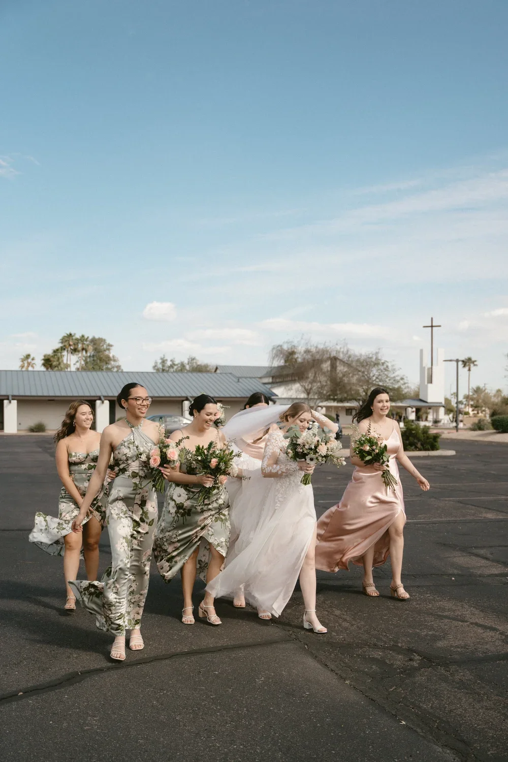 A group of women, including a bride in a white wedding dress, walking together in an outdoor parking lot. They are holding bouquets of flowers and are dressed in floral and satin dresses. The sky is blue with some clouds, and there are trees and a building in the background.