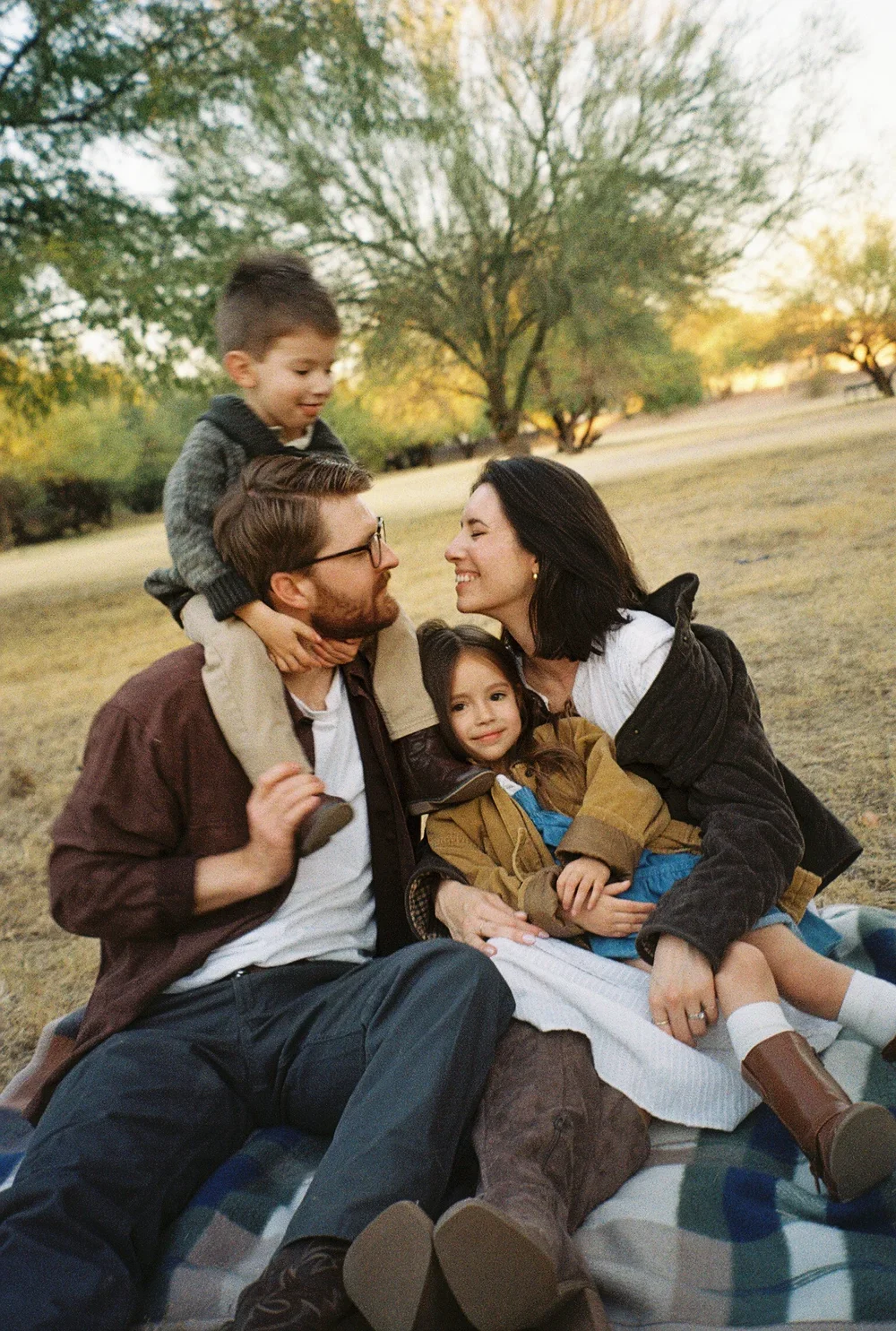 A family of four enjoying an outdoor picnic in a park during sunset. The father, mother, and two children are sitting on a blanket and interacting happily.