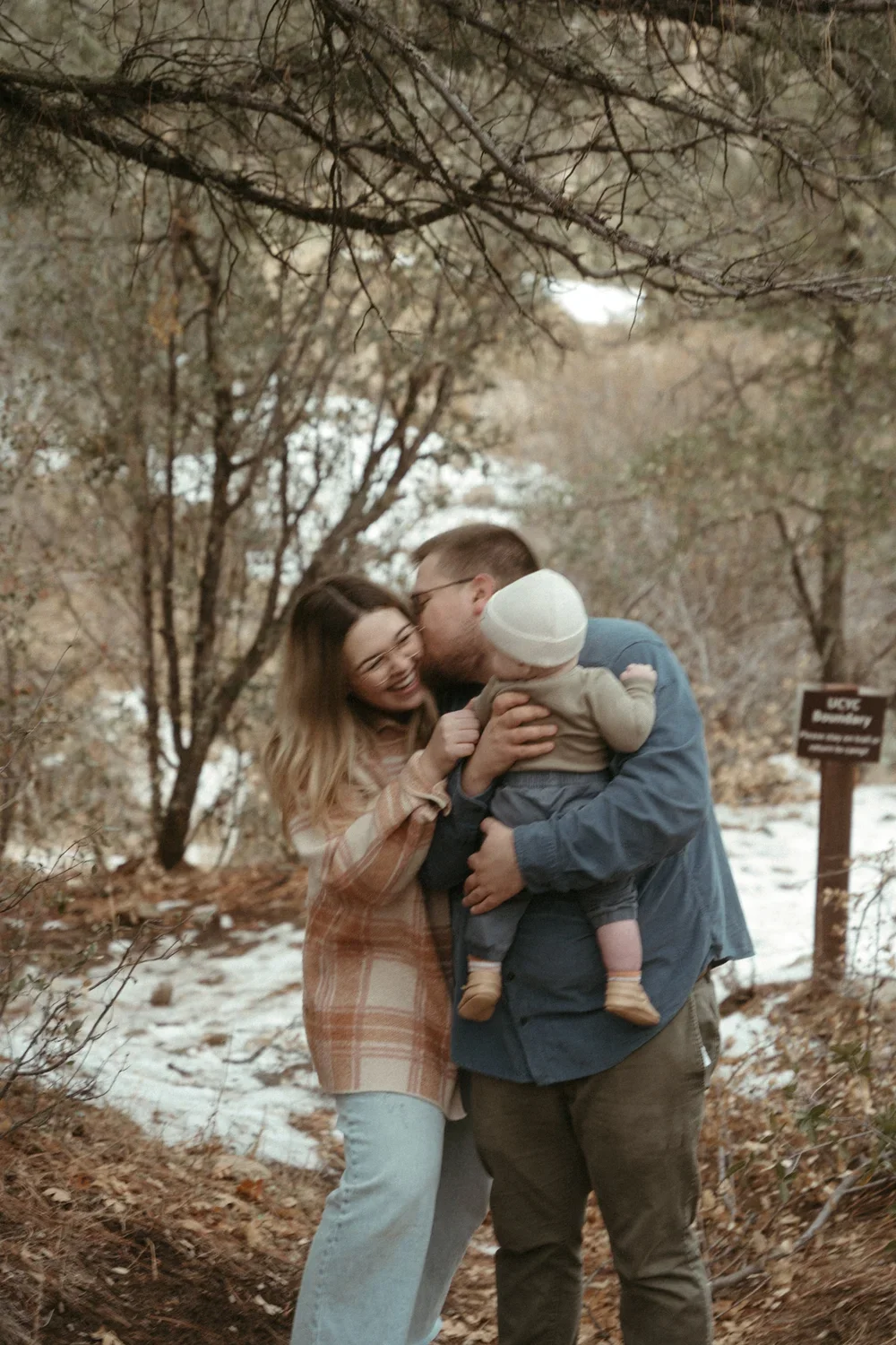 A happy family of three in a wooded outdoor setting with snow on the ground, consisting of a woman, a man, and a baby, sharing a loving moment.