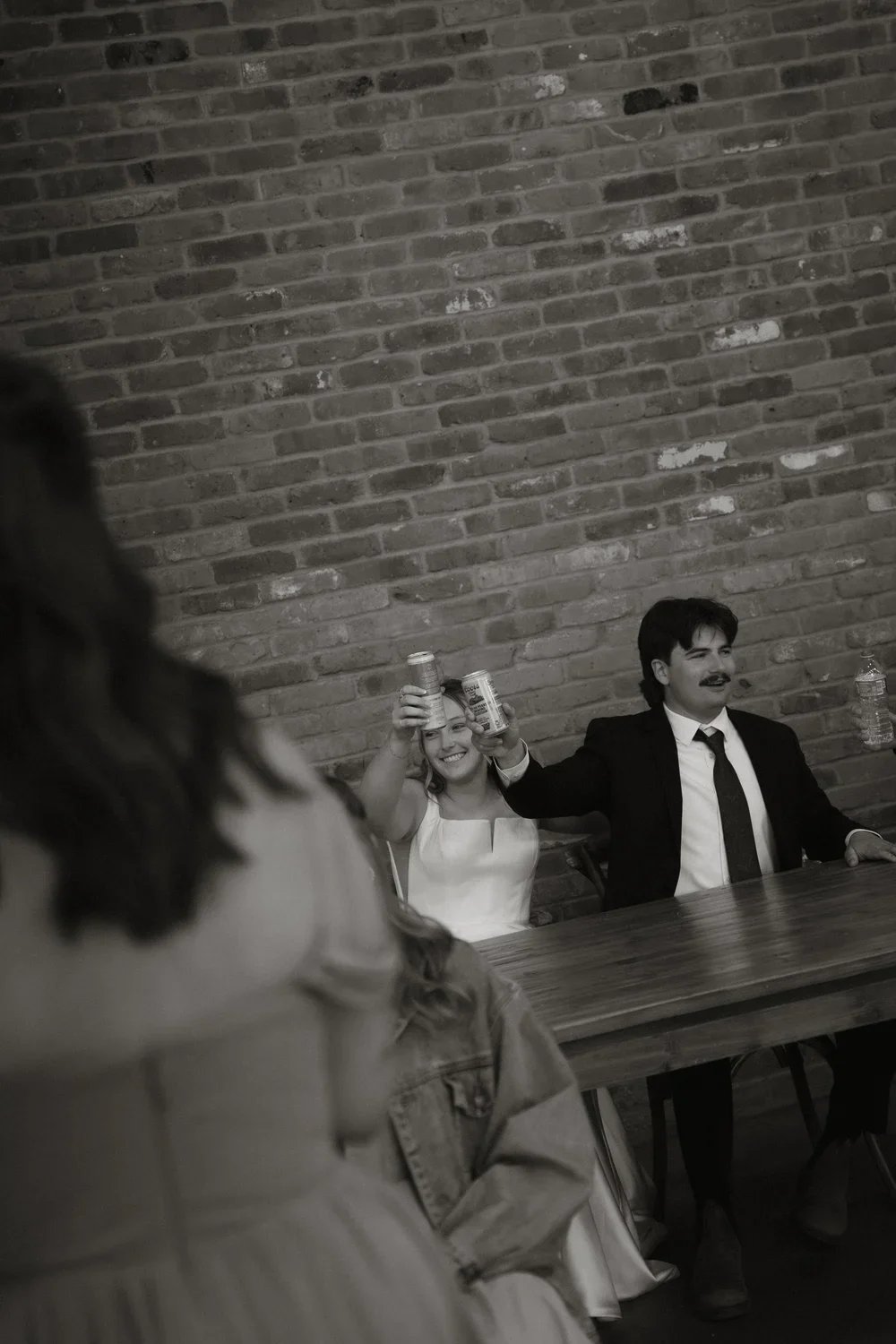 Two people sitting at a table, a woman and a man, raising cans and smiling, with a woman in the foreground partially out of focus, a brick wall in the background.