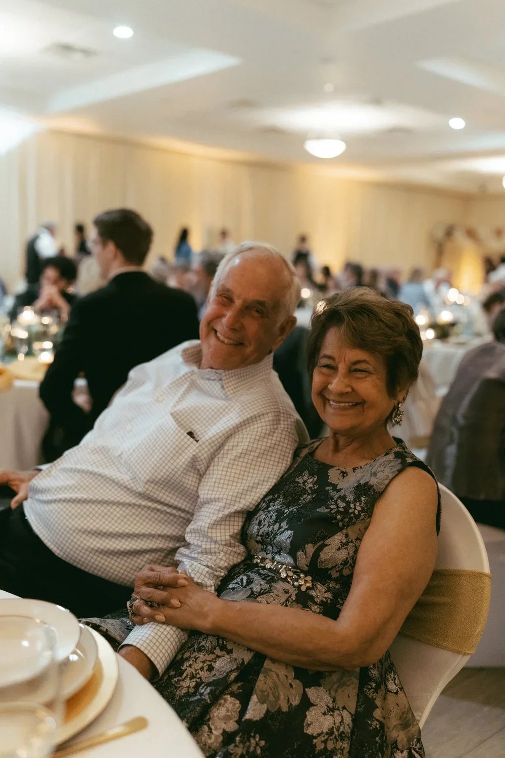 An elderly man and woman are smiling and holding hands at a formal event, sitting at a table with dinnerware, in a warmly lit banquet hall filled with other guests.