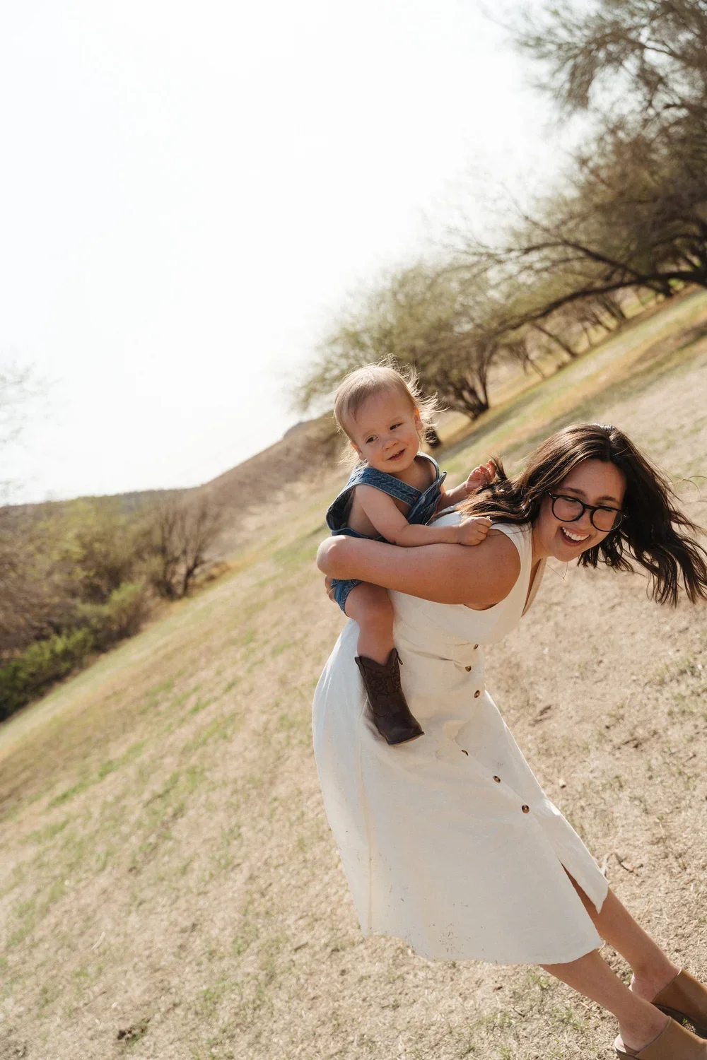 A woman with glasses in a white dress carrying a young child on her back in a dry outdoor landscape with trees and hills, both smiling.