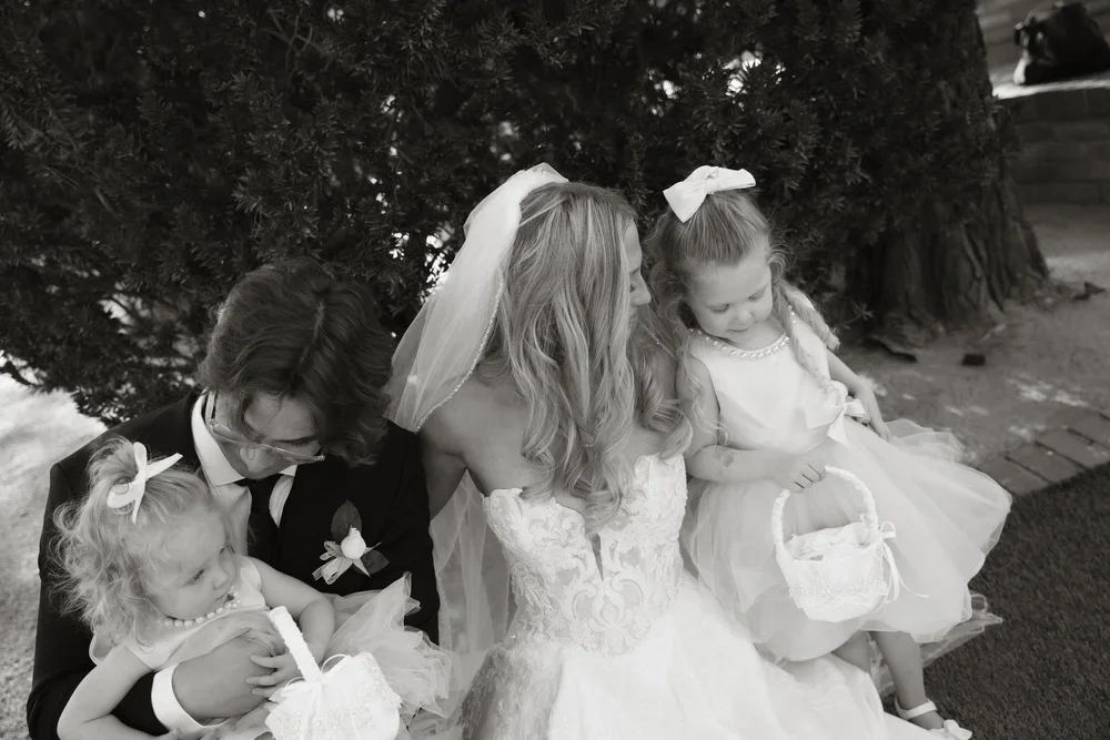 A woman in a wedding dress sitting on a bench with two young girls and a young boy, outdoors near a large tree, in black and white.