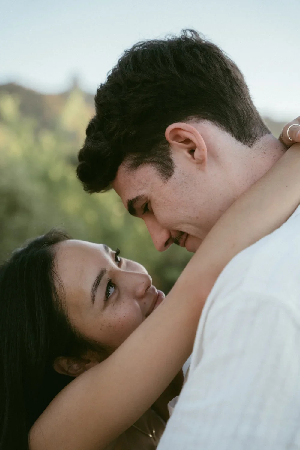 A young couple is embracing outdoors, looking into each other's eyes, with blurred greenery in the background.