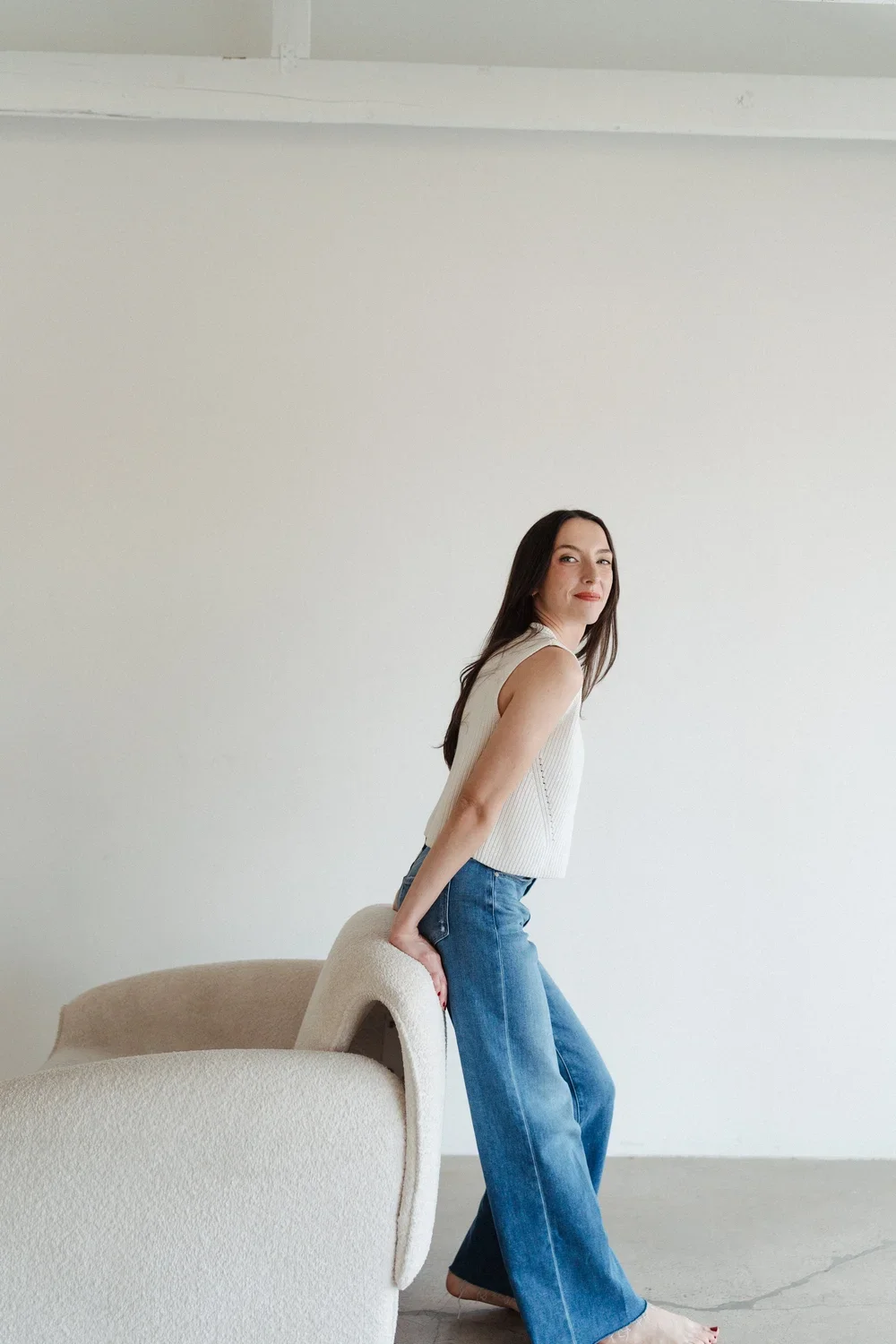 A woman with long dark hair, wearing a sleeveless white top and blue jeans, sitting backwards on a rounded beige armchair in a minimalist room with a white wall.