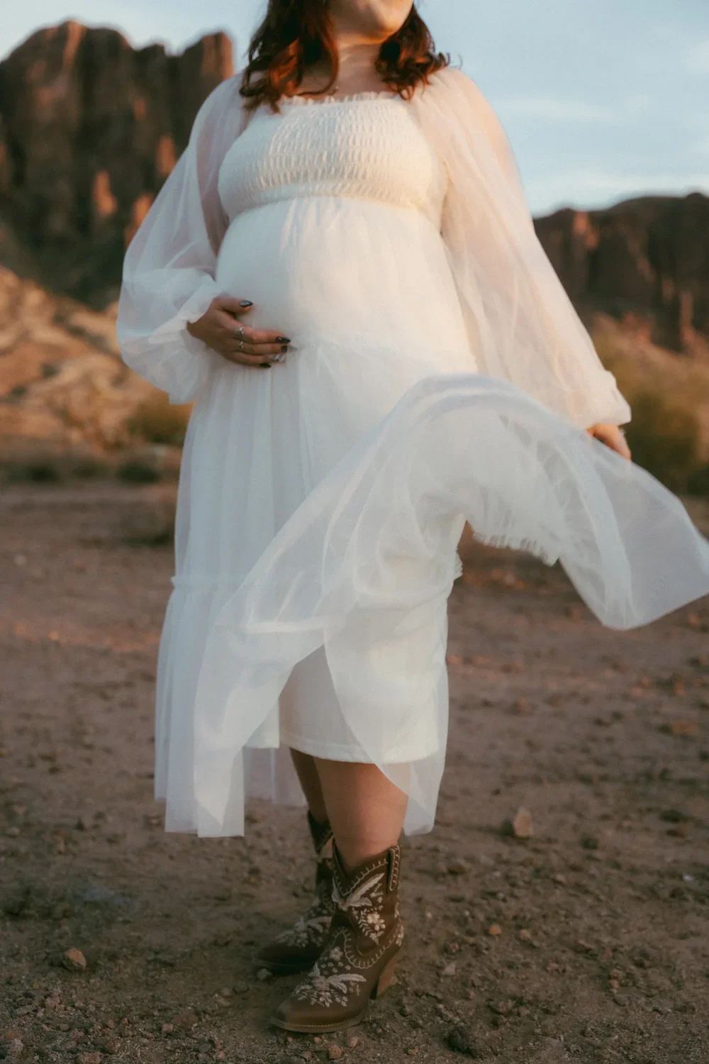 Pregnant woman wearing a white dress and cowboy boots standing outdoors in a desert landscape.