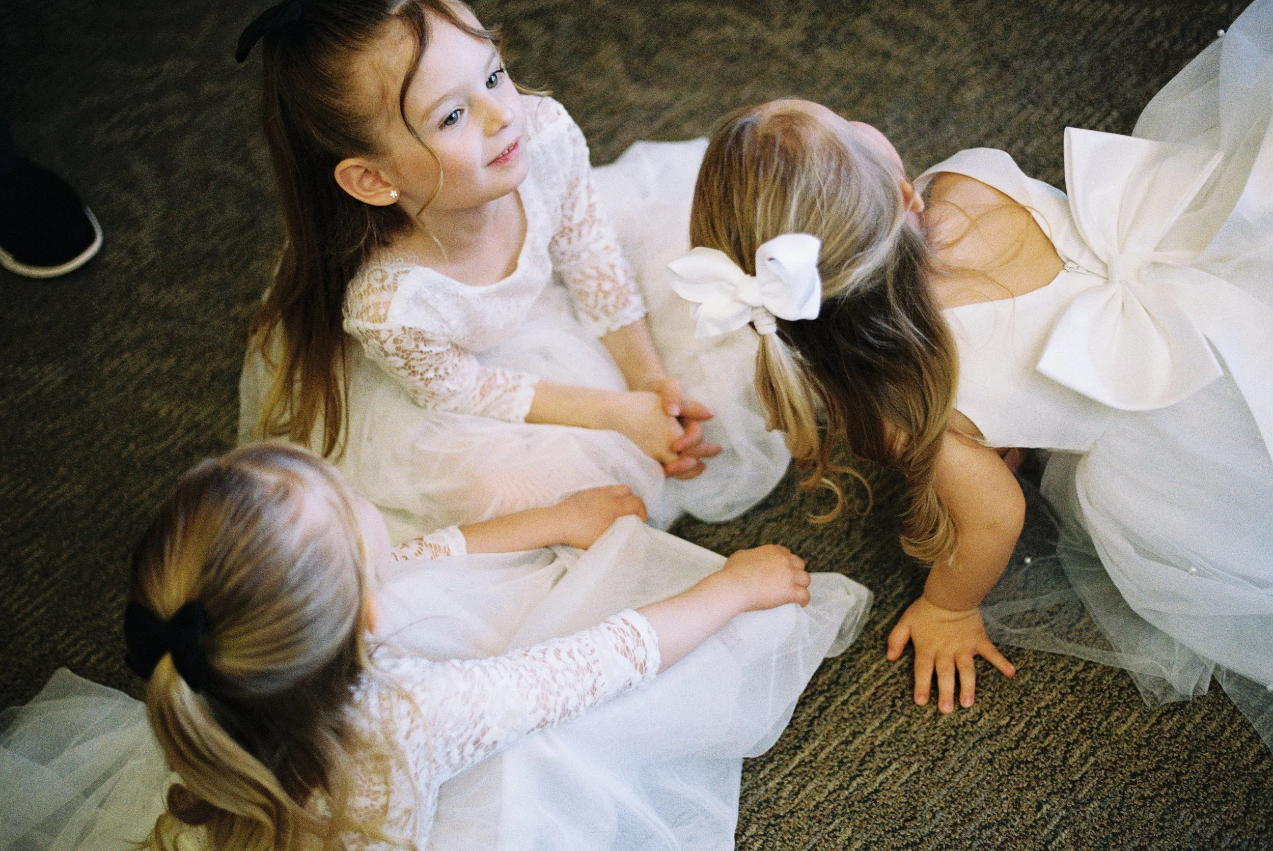 Four young girls in white dresses sitting and crouching on a carpeted floor, with two of them wearing bows in their hair.