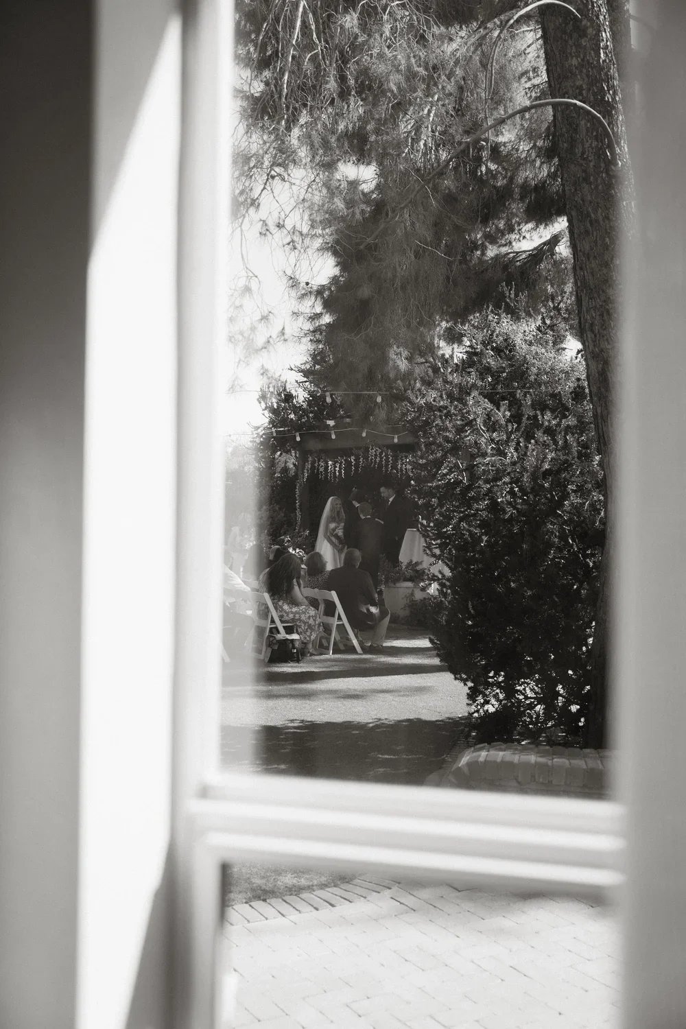 Sight of a wedding ceremony through a window, with people seated outside, trees, and a couple standing at the altar.