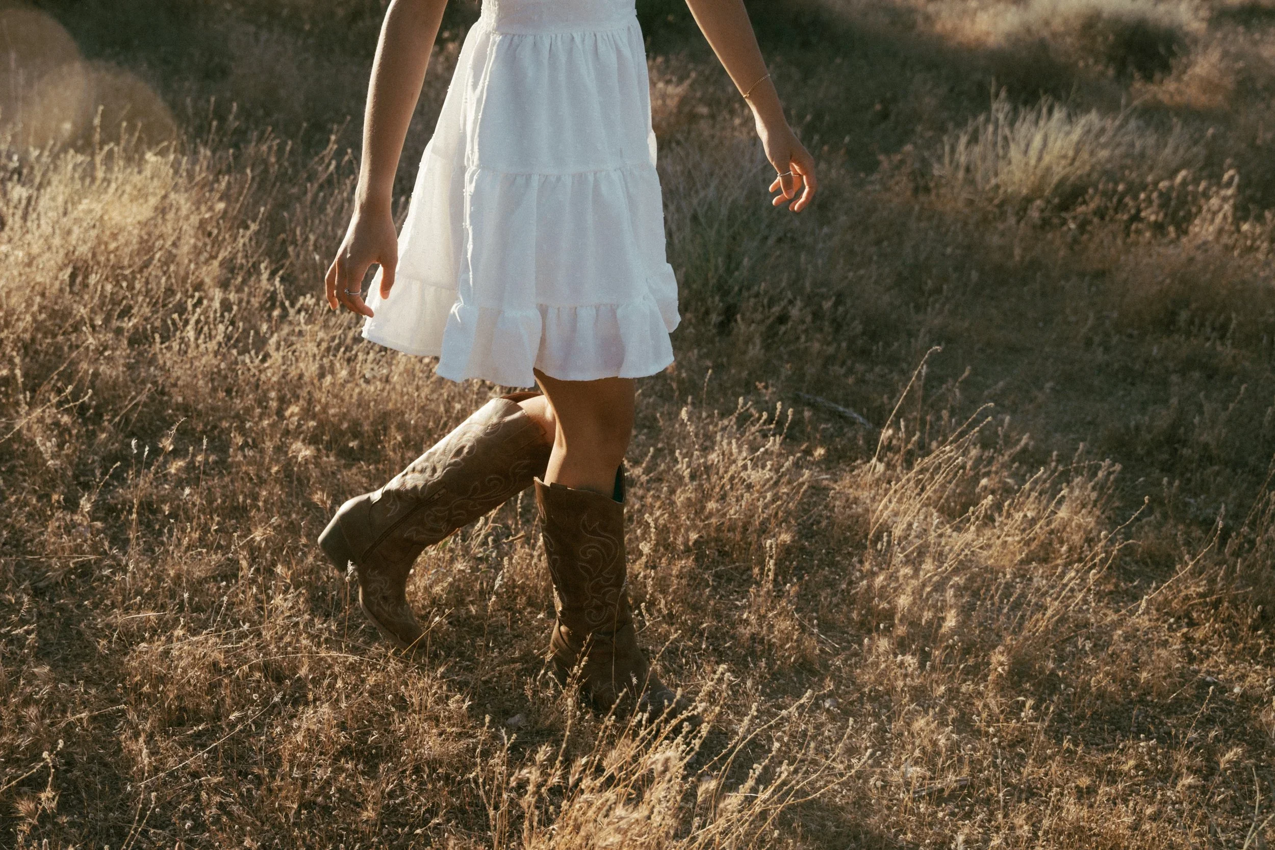 A woman walking through a dry field wearing a white dress and cowboy boots, with the sunlight creating a warm glow.