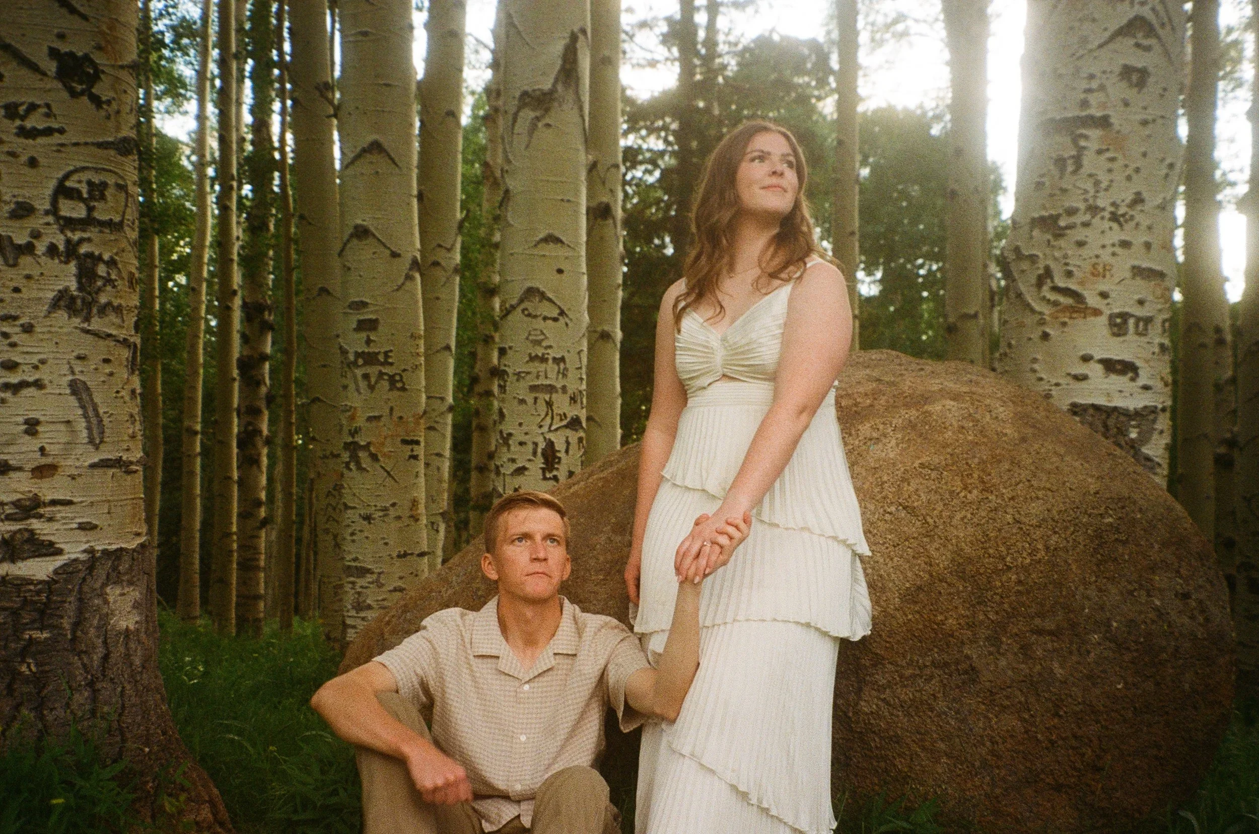 Three people in a forest setting with aspen trees. A woman in a white dress stands on a rock holding hands with a seated man in a beige shirt, while another man in a plaid shirt looks on from lower in the scene.