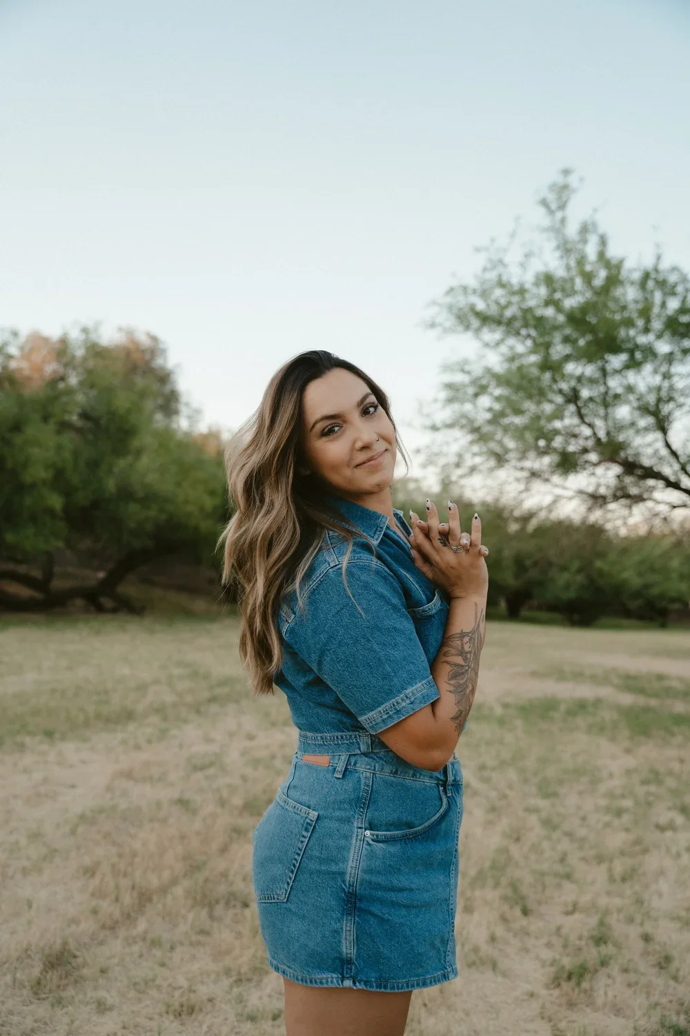 A woman with long wavy hair, wearing a denim shirt and skirt, standing outdoors in a grassy field with trees, during early evening or late afternoon.